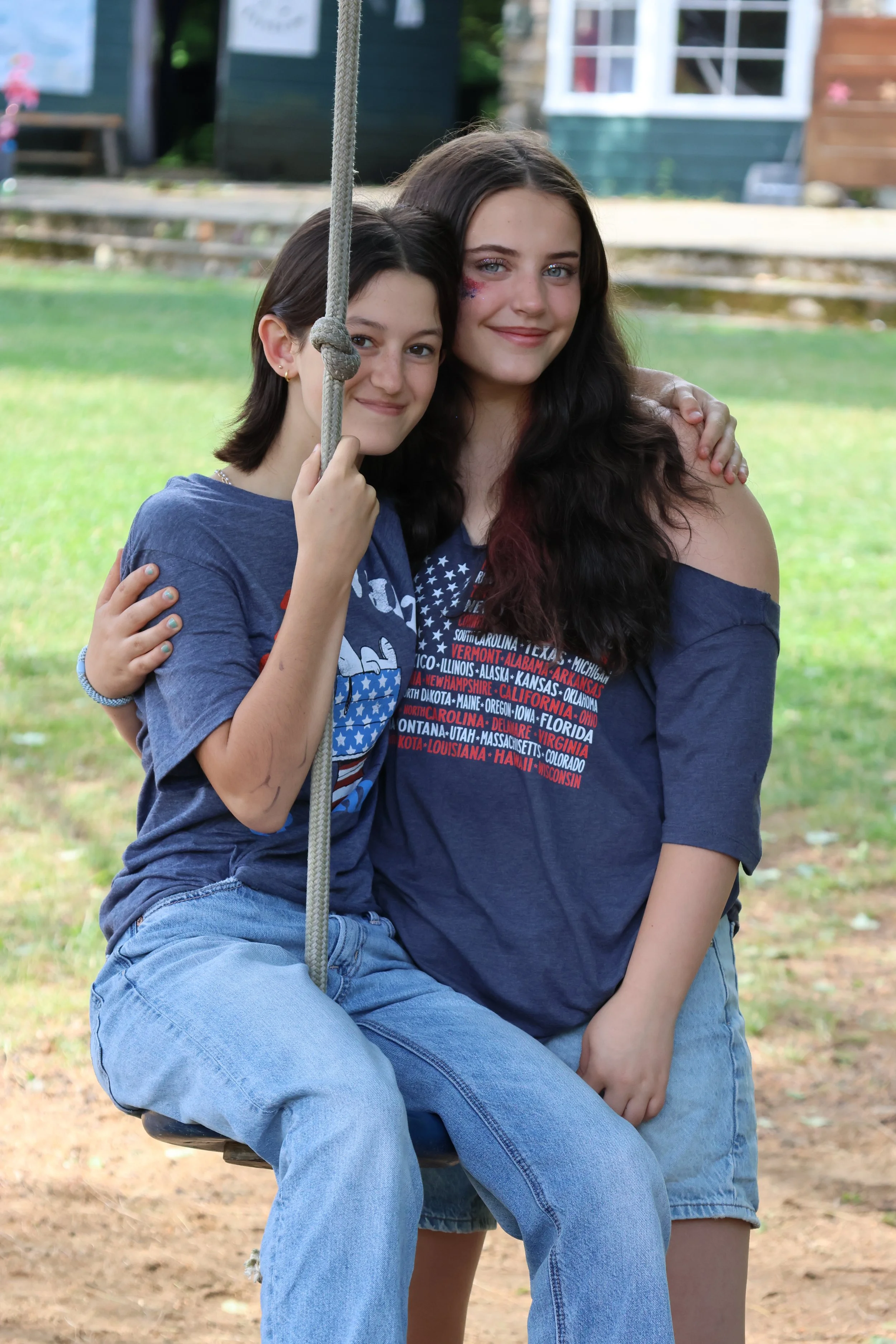 Two young women sitting on a swing outdoors, smiling at the camera, with a house and grass in the background.