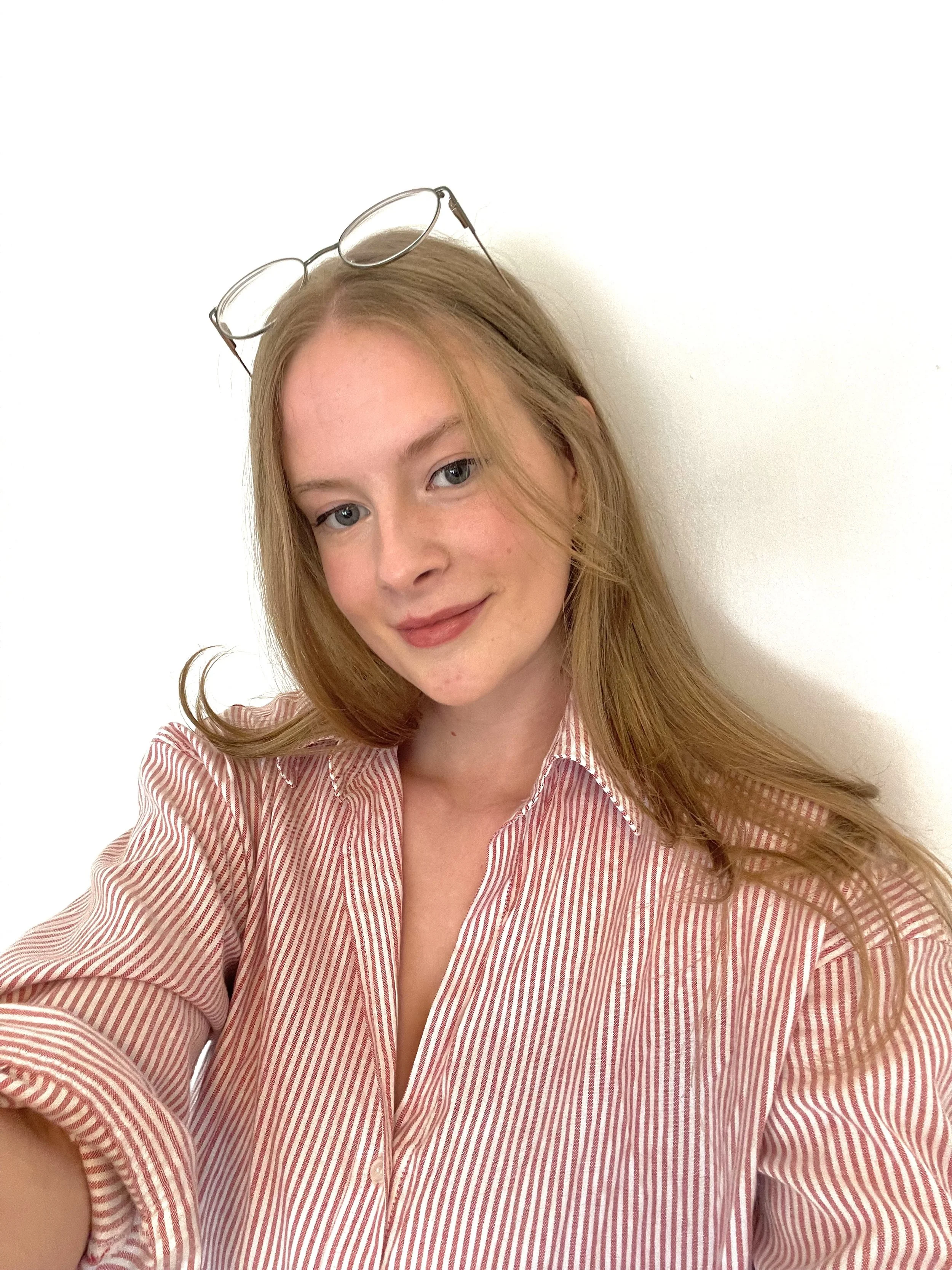 A young woman with long reddish hair wearing a pink and white striped shirt and glasses on her head, smiling against a plain white background.