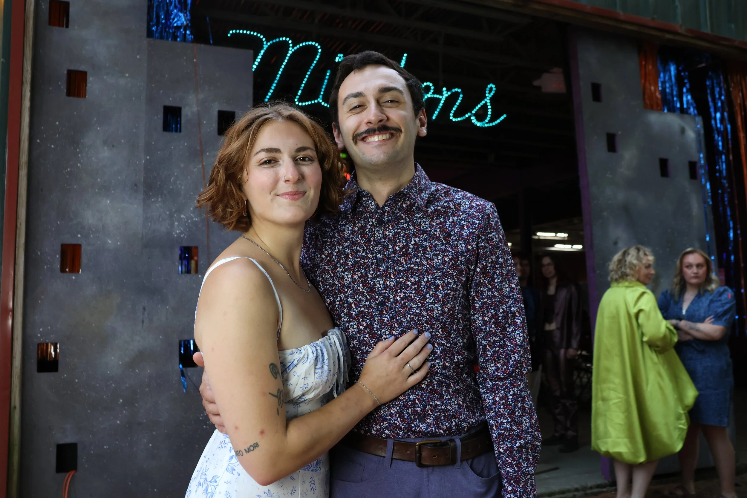 A smiling woman and man standing close together at an indoor event, with a neon sign in the background that says "Motions". The woman has light skin, curly red hair, and is wearing a dress with a thin strap, showing tattoos on her arm. The man has li