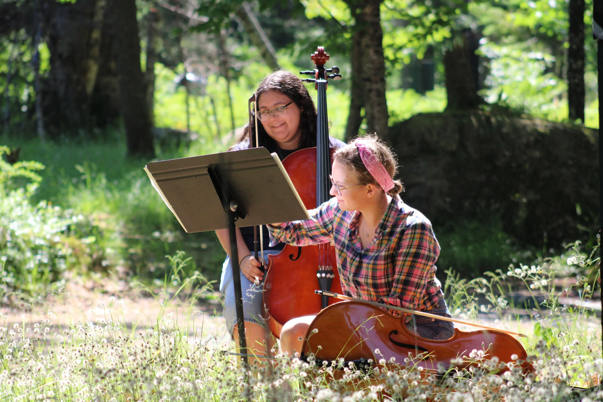 Two women are outdoors in a forest, one playing the violin and the other playing the cello, with sheet music on a music stand in front of them.