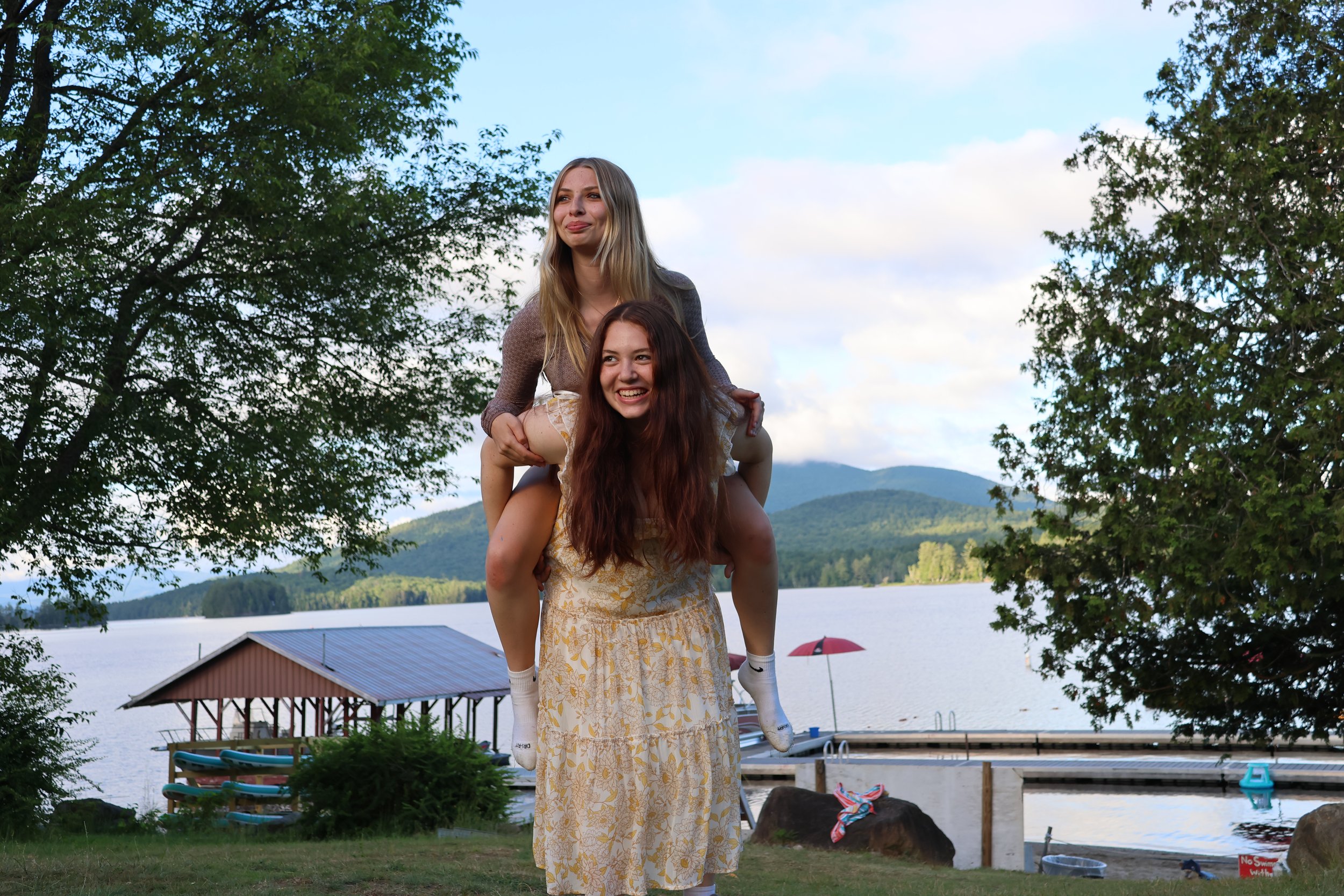 Two young women are outdoors by a lake with mountains in the background; one is giving the other a piggyback ride, both smiling and enjoying the scene, surrounded by trees and some docked boats.