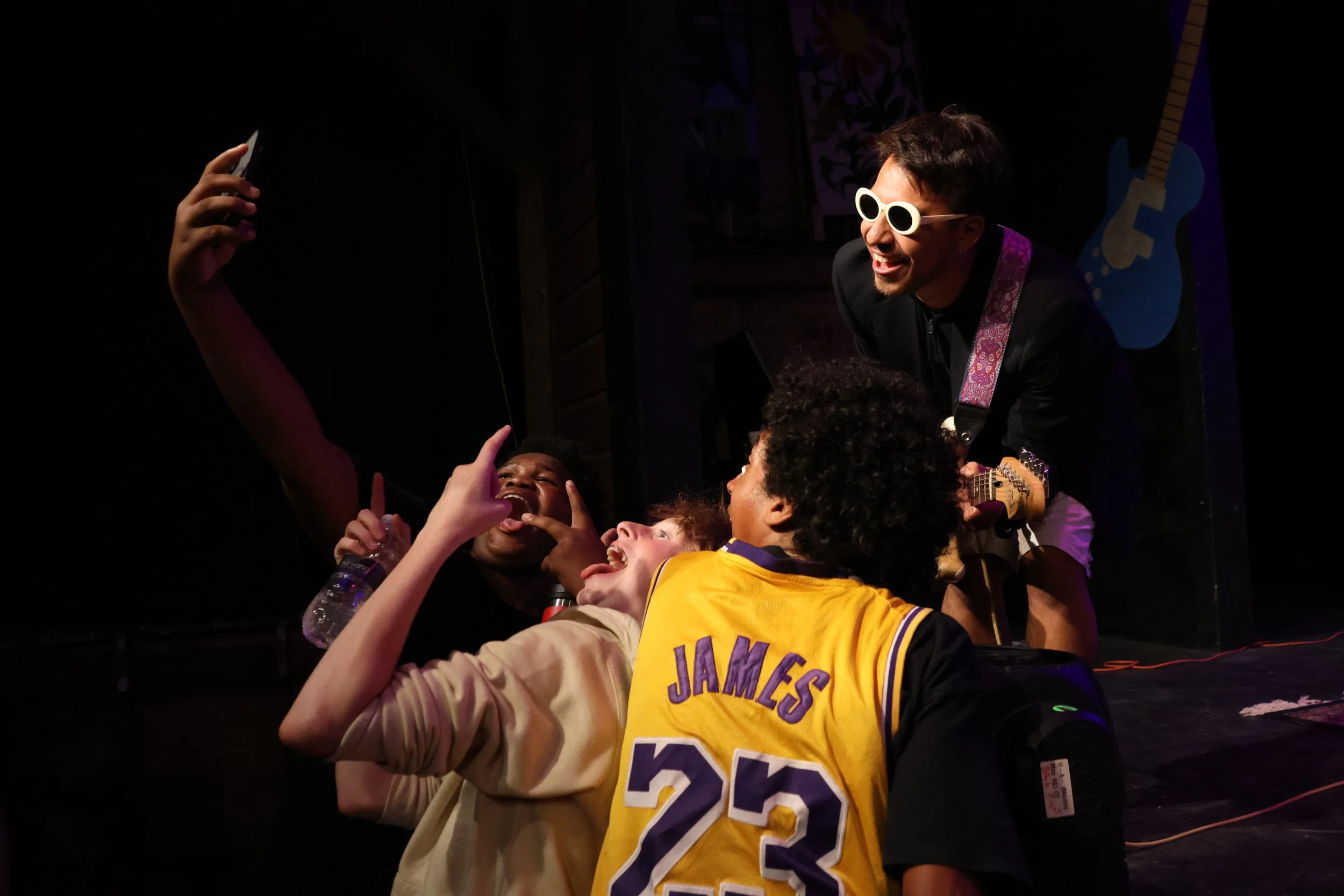 A performer with white sunglasses and curly hair is singing and playing guitar on stage, surrounded by young fans, some taking photos, in a lively concert scene.
