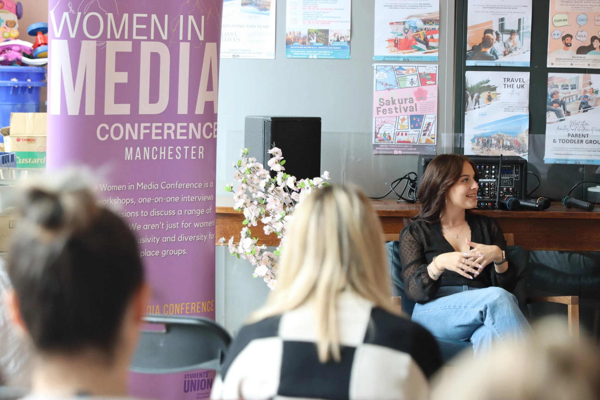 A woman with shoulder-length brown hair, wearing a black top with sheer sleeves, sitting and smiling at a conference or event. There are several posters on the wall behind her and she appears to be engaged in conversation. The background includes a jewelry-like audio equipment with microphones and a large pink-purple banner for the 'Women in Media Conference Manchester.' The foreground includes blurred heads of other attendees.