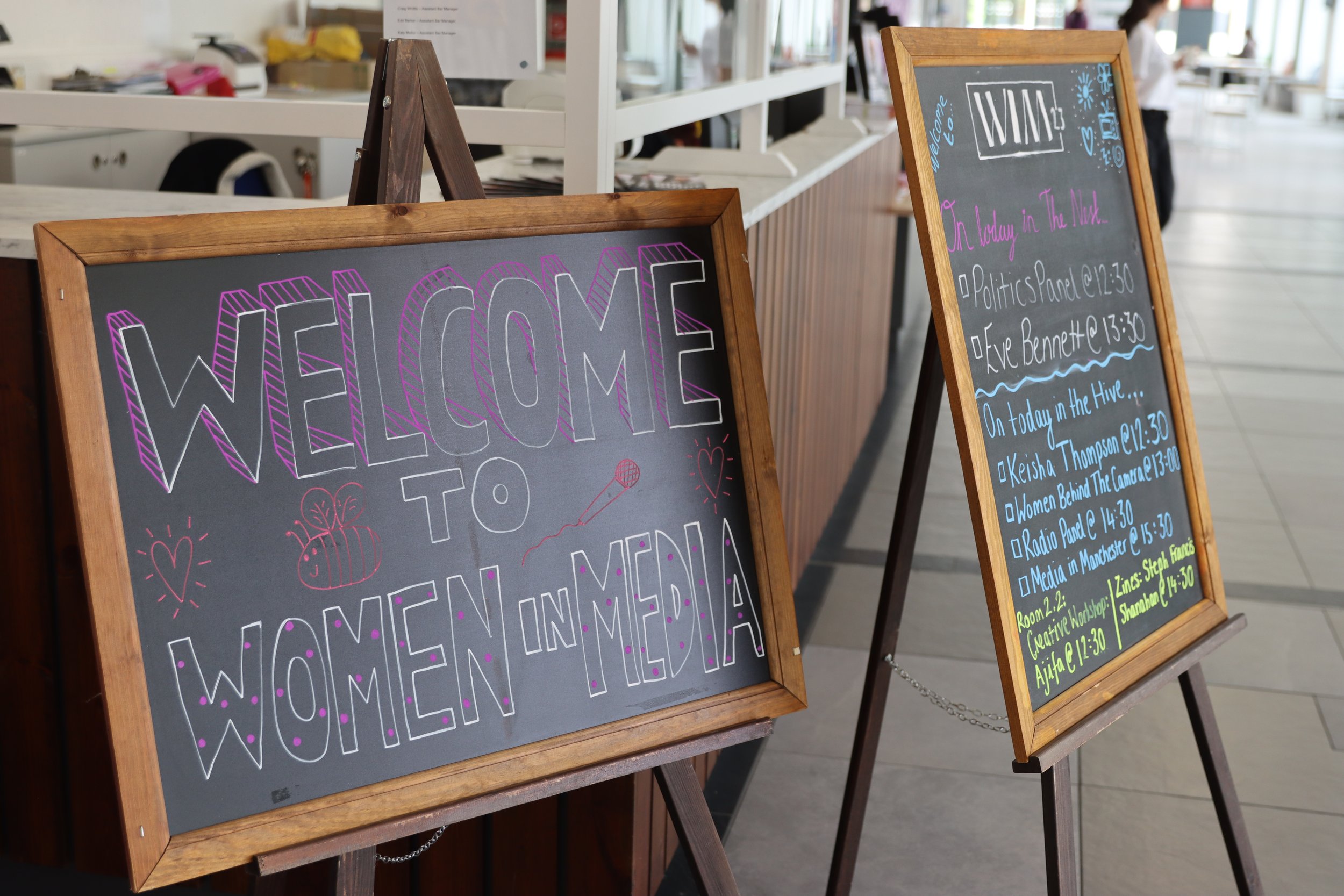Two large chalkboards on wooden easels, one with colorful writing that says "Welcome to" and festive drawings, the other with event schedule details handwritten with different colors in an indoor setting with people and a reception desk in the backgr