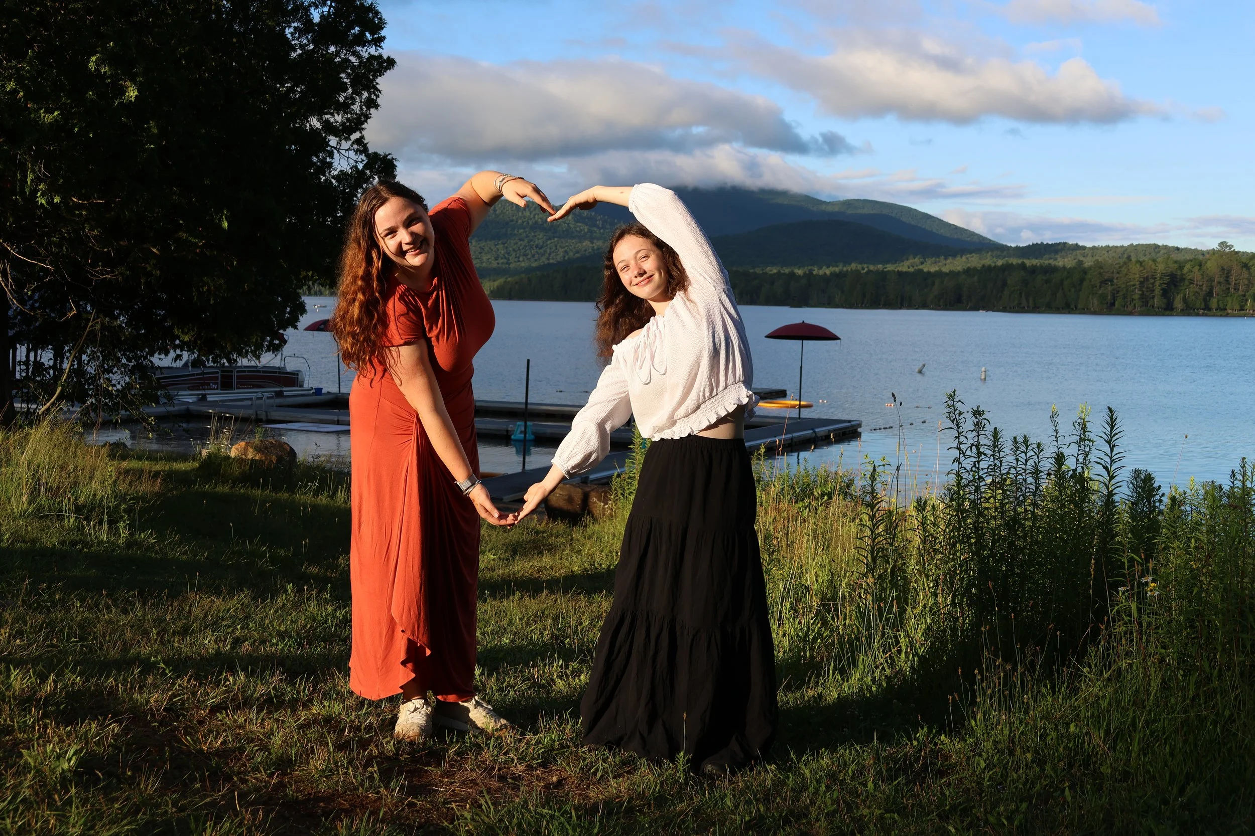 Two women standing outdoors near a lake, forming a heart shape with their arms and hands, with mountains and trees in the background.