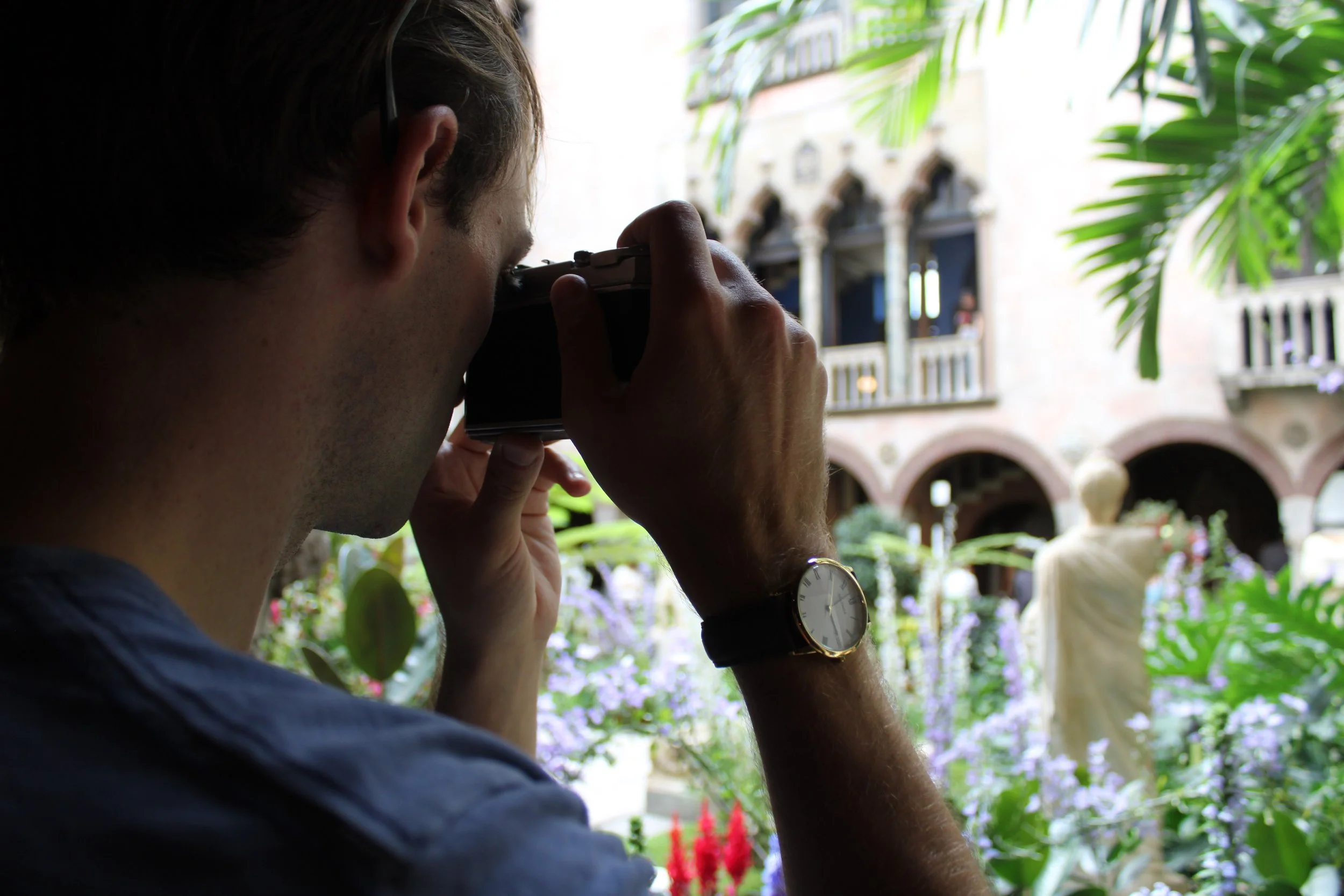 A man is looking through a camera, capturing a photo in an outdoor setting filled with greenery and decorative architecture.