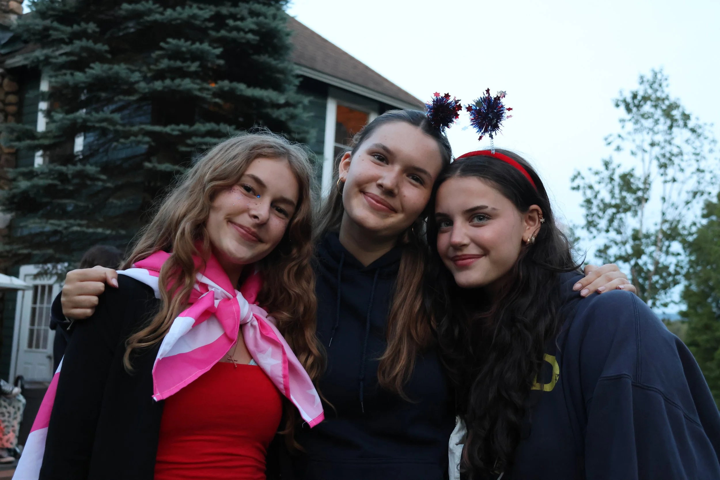 Three young women standing close together outdoors, smiling, with a house, trees, and a cloudy sky in the background. Two women wear festive headbands with red, white, and blue decorations.