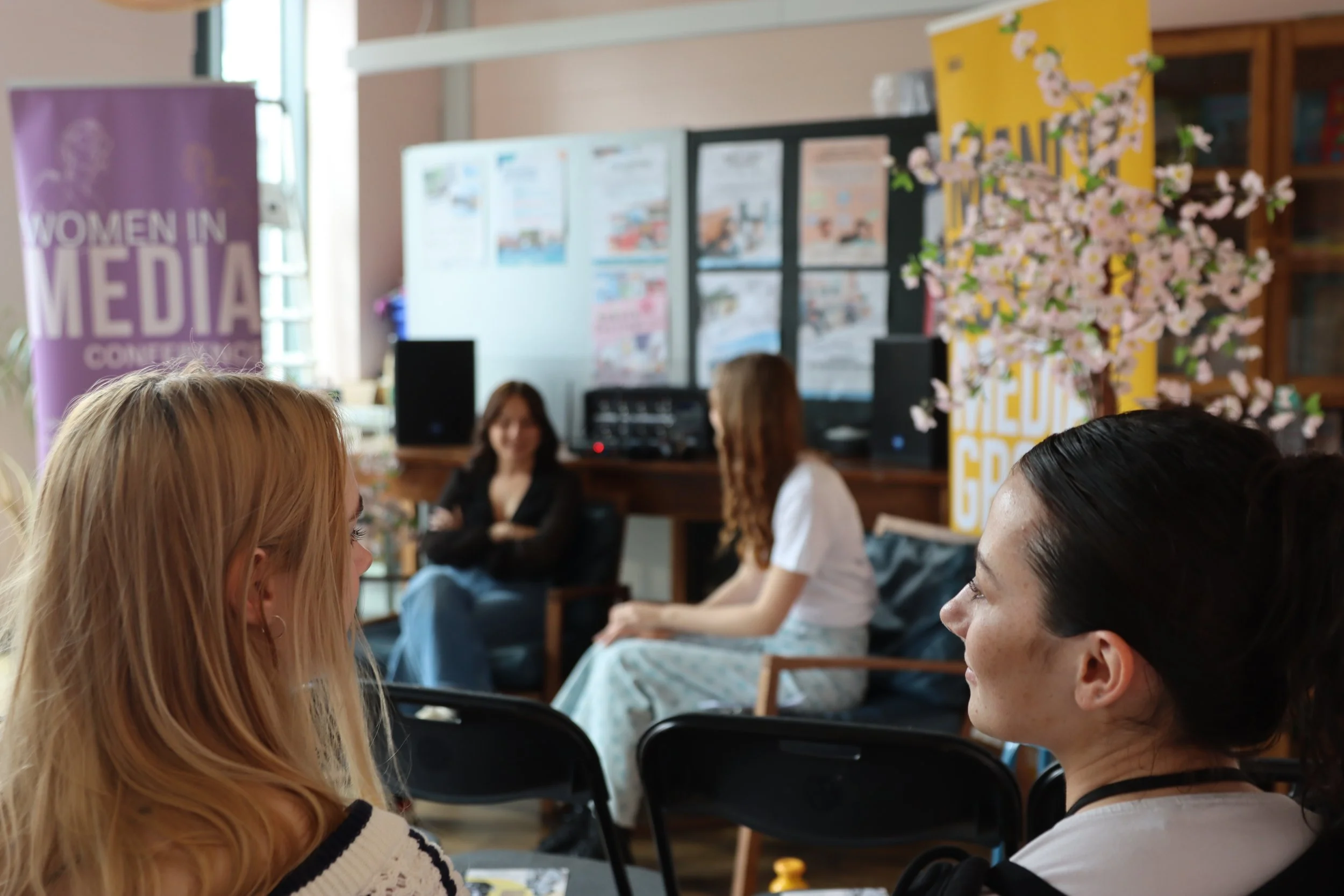 People sitting and talking in a community center with informational posters on the wall and a large decorative cherry blossom tree.