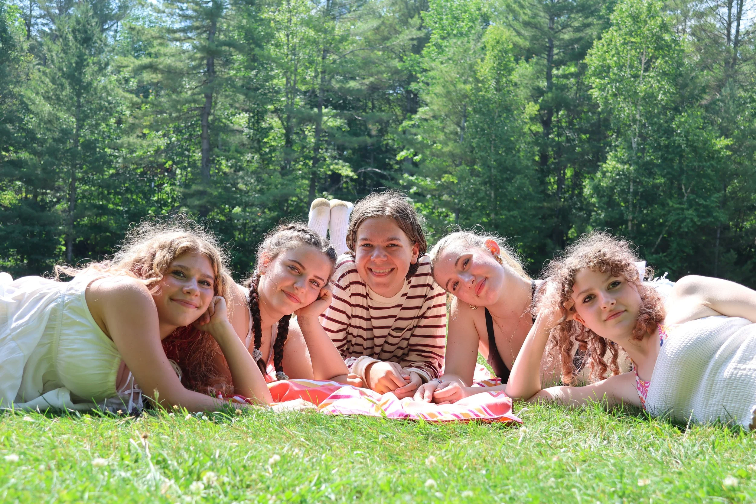 Group of six young women lying on grass in a park during daytime, smiling and posing for the camera with trees in the background.