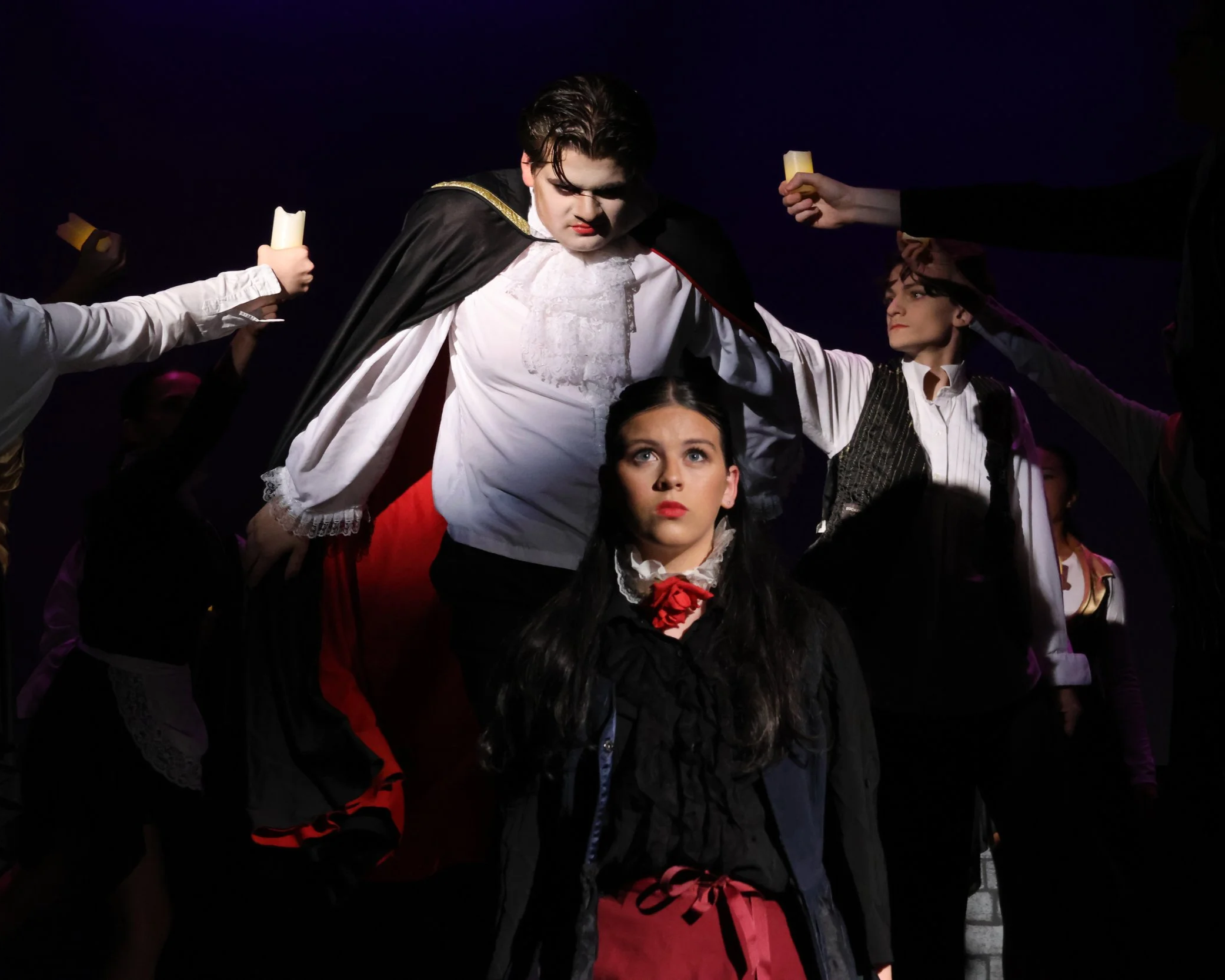 A scene from a theatrical performance with a person dressed as Dracula, wearing a black cape with a gold trim and a white lace shirt, surrounded by other performers in period costumes, some holding candles, against a dark background.