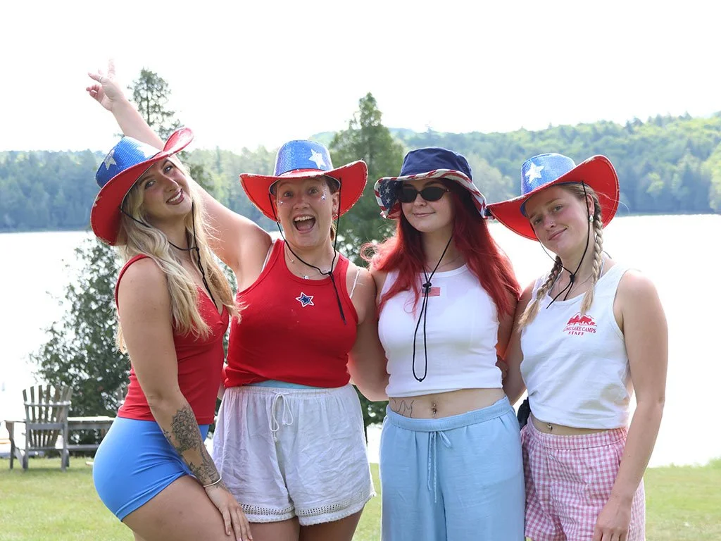 Four young women wearing patriotic hats and summer clothes standing outdoors near a lake on a sunny day.