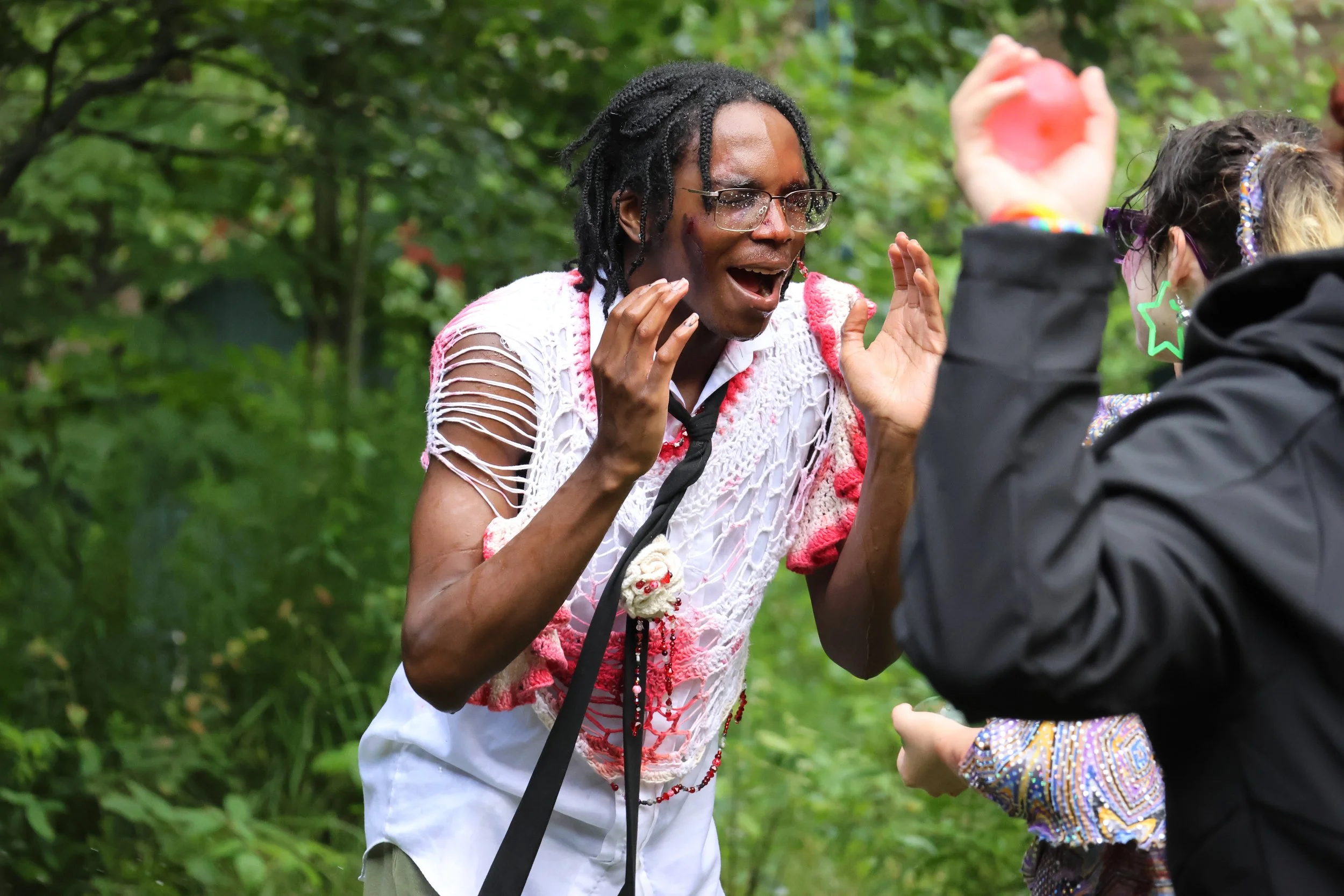 A person wearing glasses and a white shirt with pink details, with dreadlocks, appears to be surprised or excited while interacting outdoors with another person who is partially visible, wearing glasses, star-shaped earrings, and colorful clothing, i