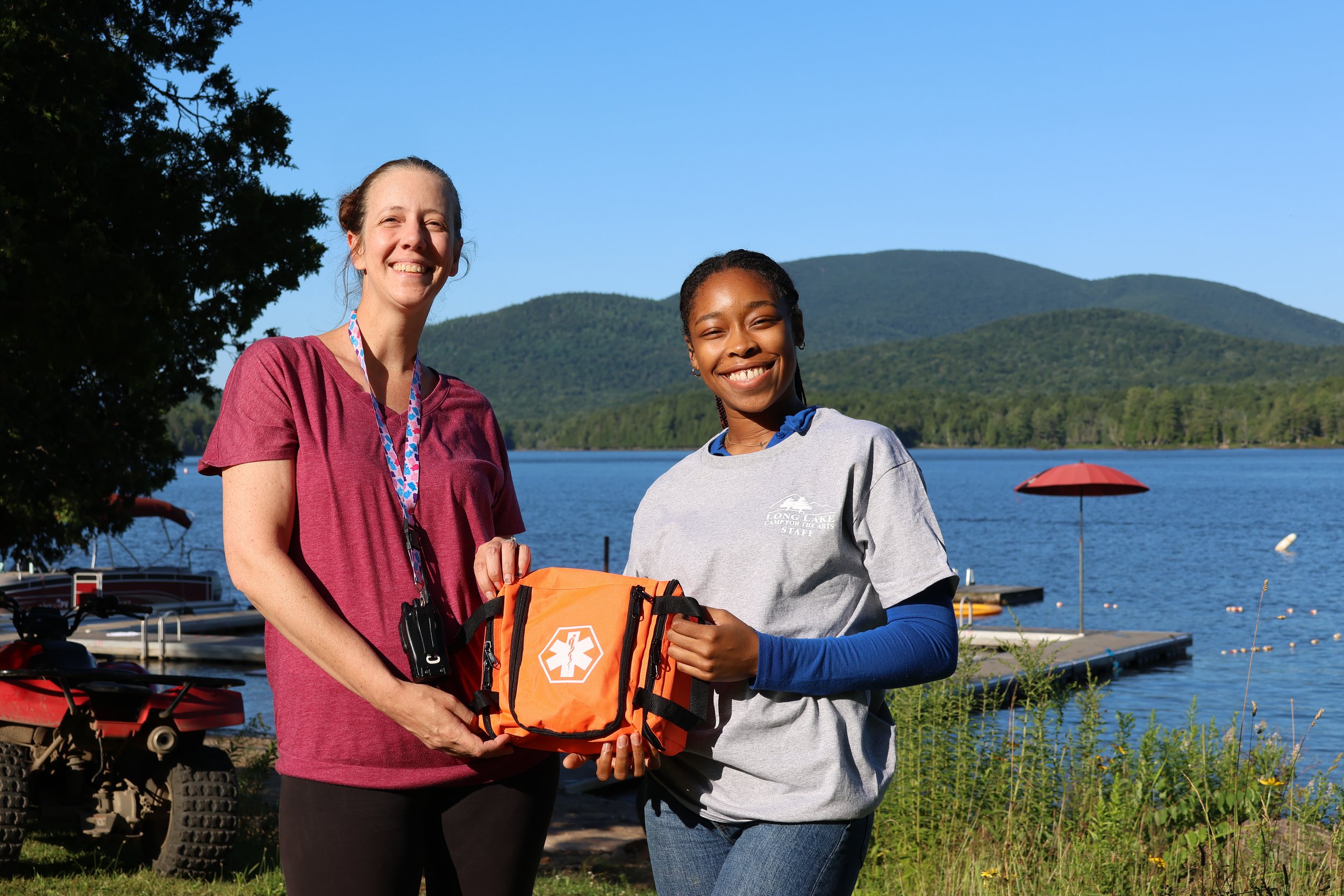 Two women smiling in front of a lake with green mountains in the background, holding an emergency medical kit.