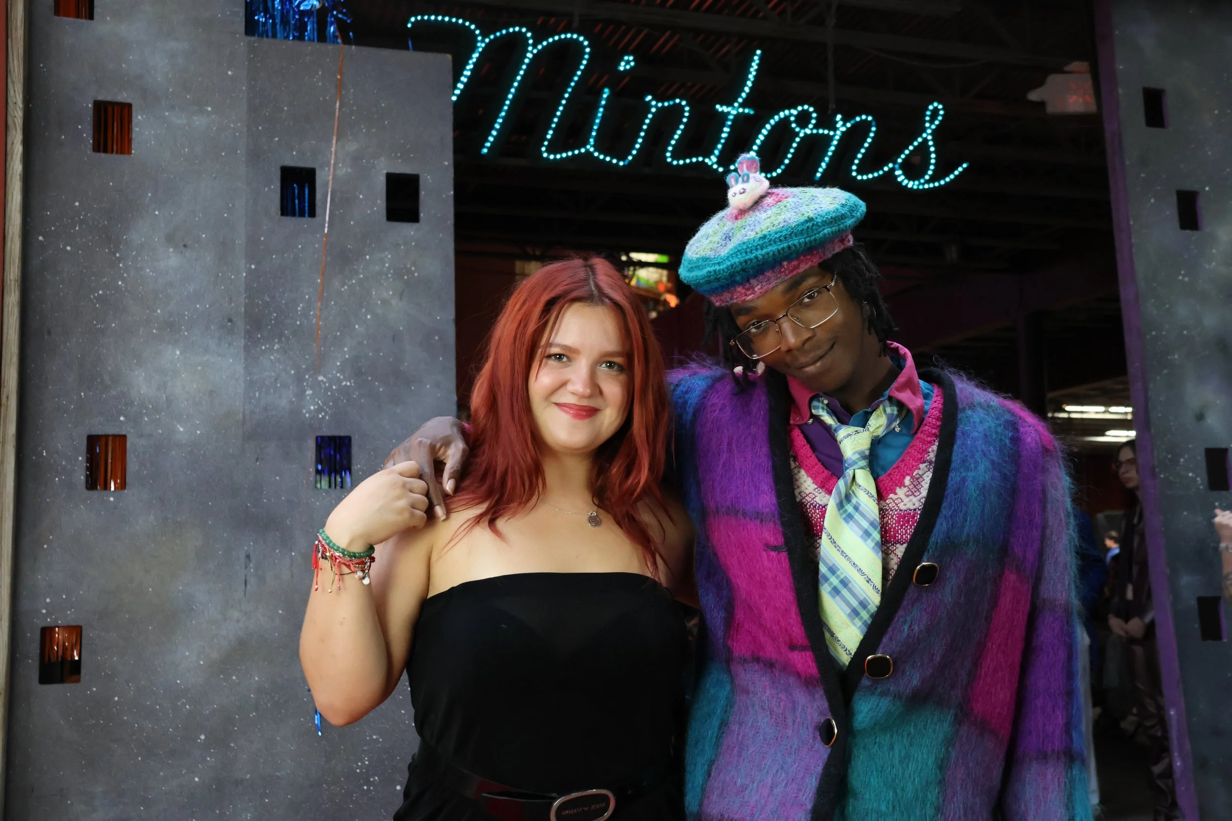 A young woman with red hair and wearing a black strapless top, smiling and posing with an older man wearing glasses, a colorful knitted hat with a unicorn, and a vibrant multicolored jacket. They are standing in front of a decorative background with 