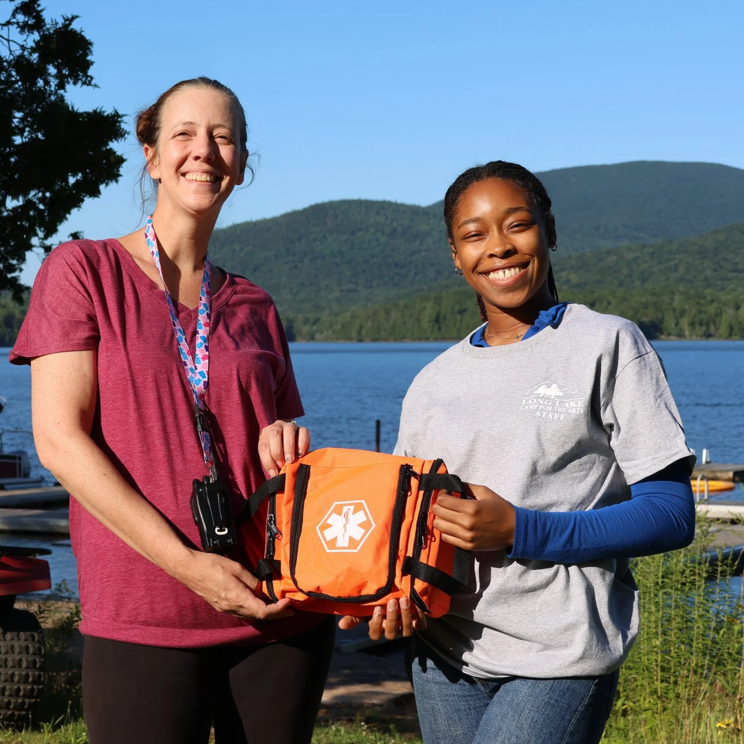 Two women smiling outdoors near a lake with mountains in the background, one holding an orange emergency medical kit.