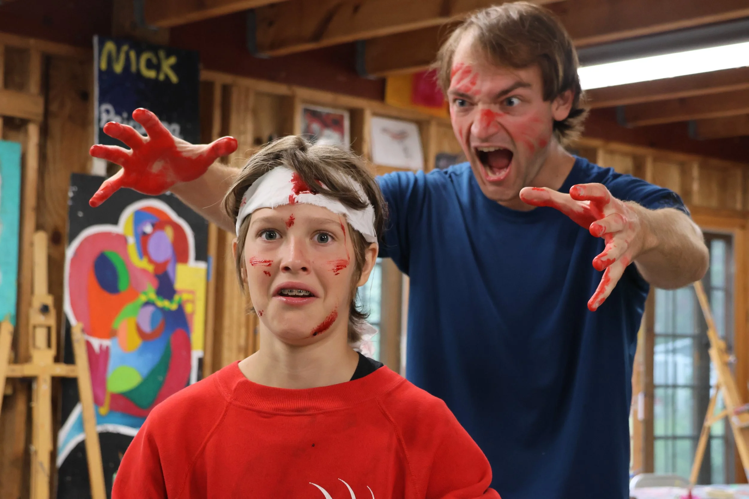 A man with paint on his face and hands is yelling and reaching towards a young boy with paint on his face, all inside a wooden art studio with paintings in the background.