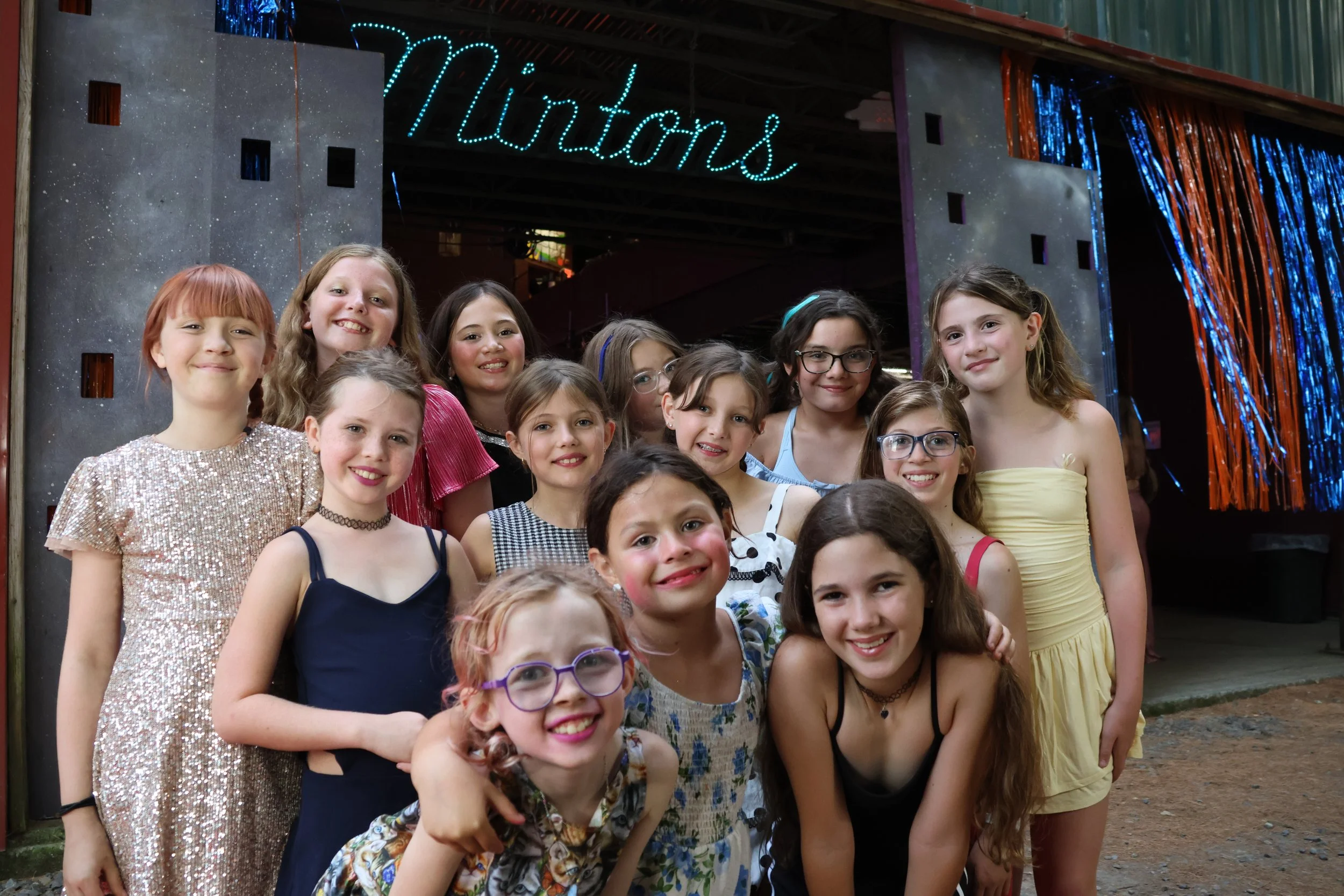 Group of smiling young girls gathered together at an event with a sign that says 'Vortex' in neon lights behind them.
