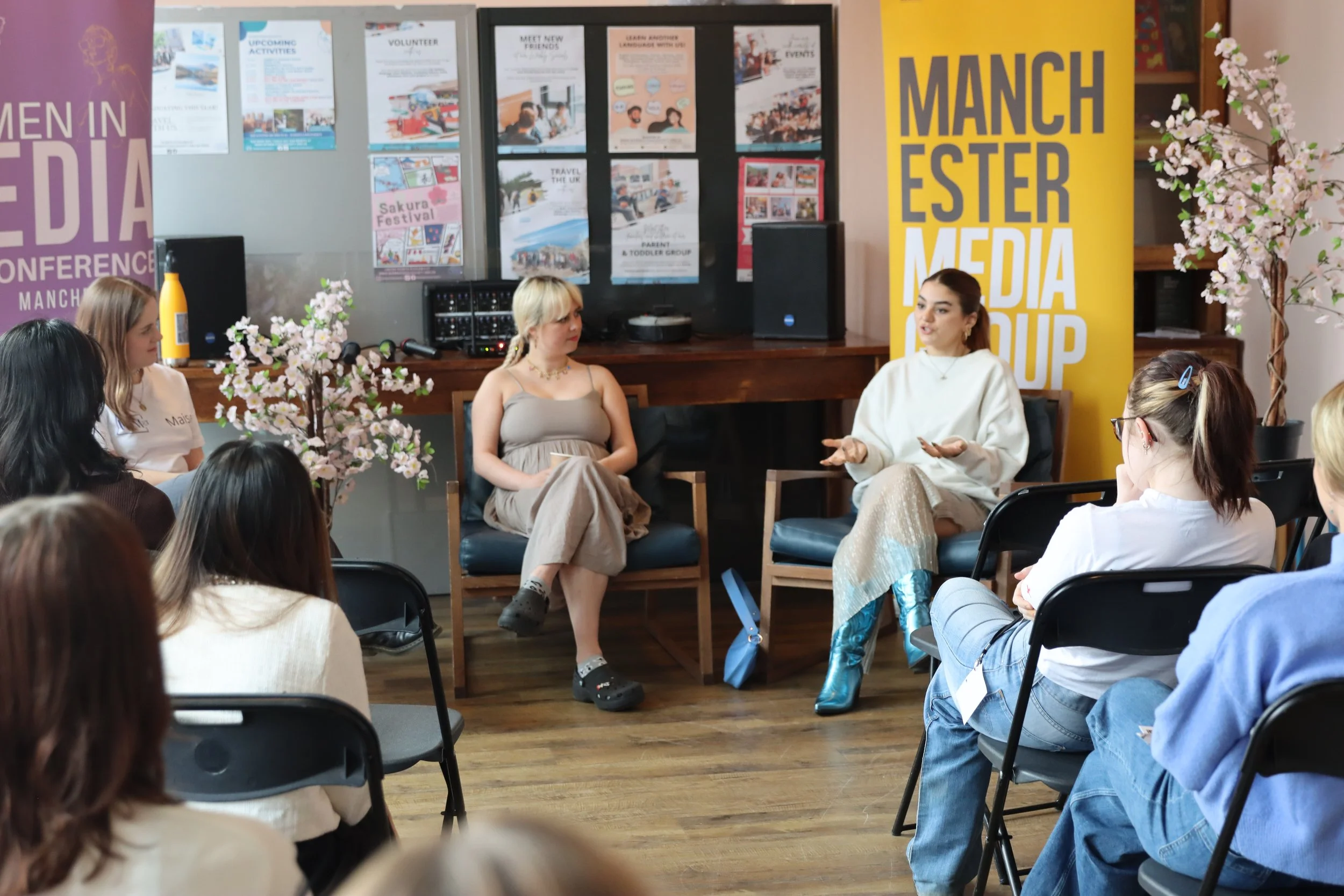 A panel discussion or interview taking place in a conference room with multiple women seated in chairs. The room is decorated with pink cherry blossom trees and colorful posters on the walls, including bright yellow and purple banners, indicating a m