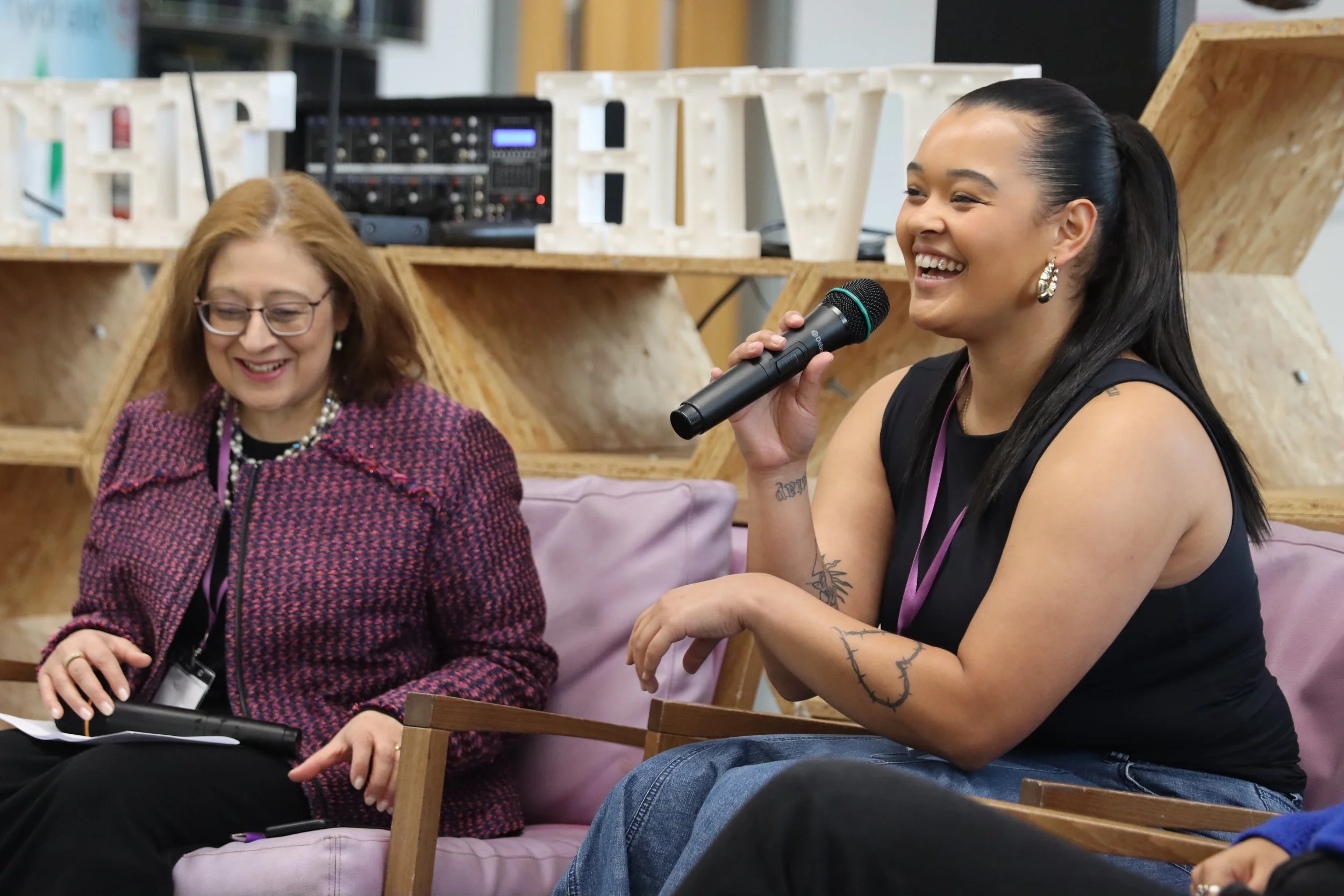 Two women sitting on a pink couch, one with red hair and glasses, the other with dark hair in a ponytail, speaking into a microphone, at an event with decorative large letters in the background.