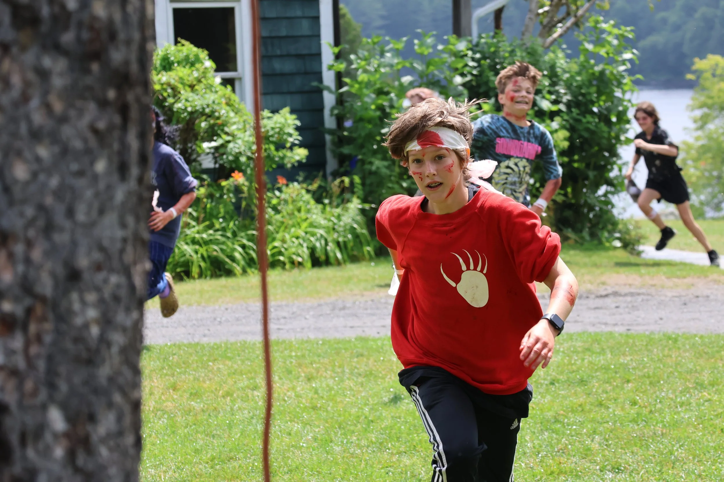 Children participating in an outdoor activity, with a boy in a red shirt running and another boy behind him with face paint, near a tree and greenery, during daytime.