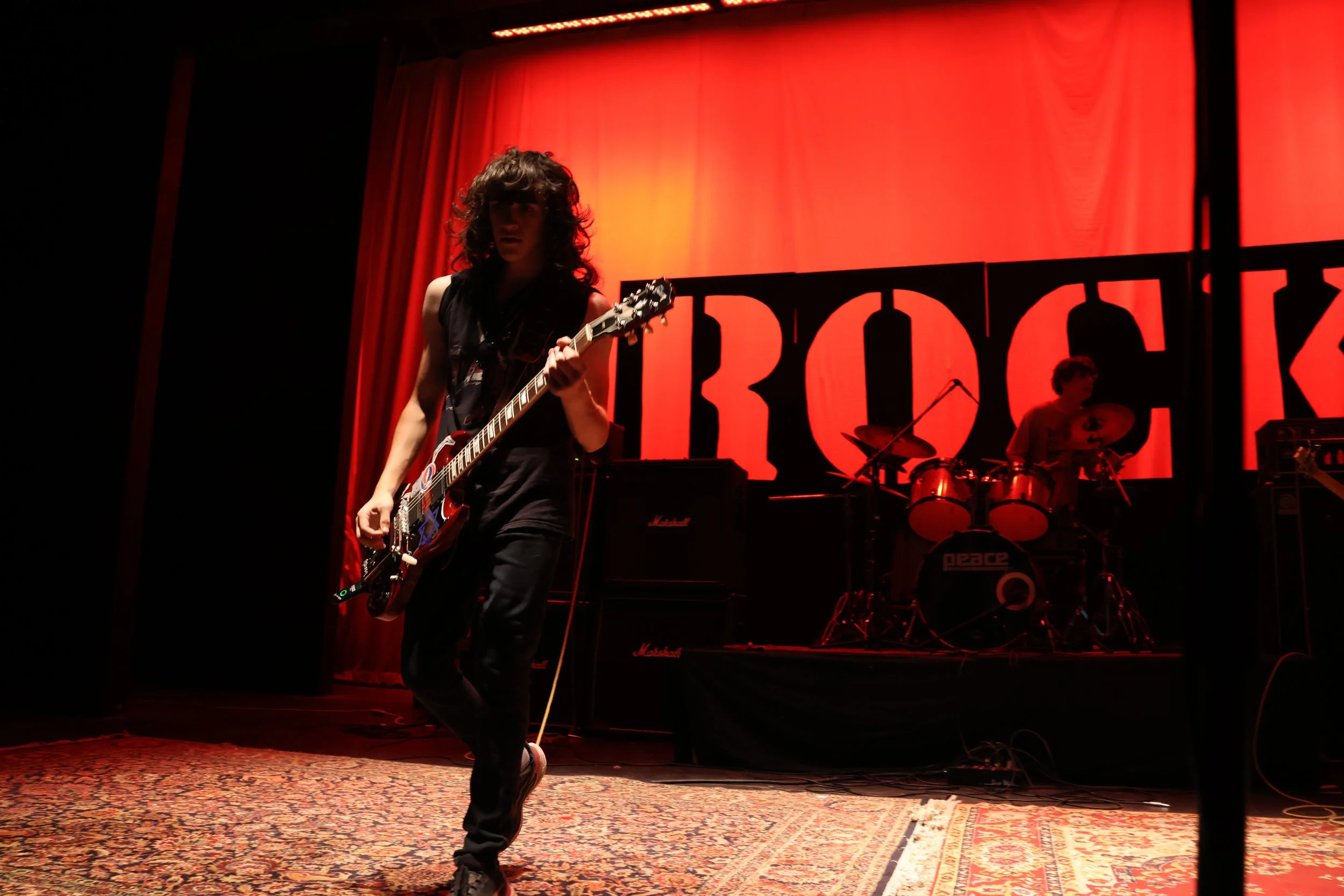 Musician with curly hair playing electric guitar on stage with red backdrop and drum set in background.