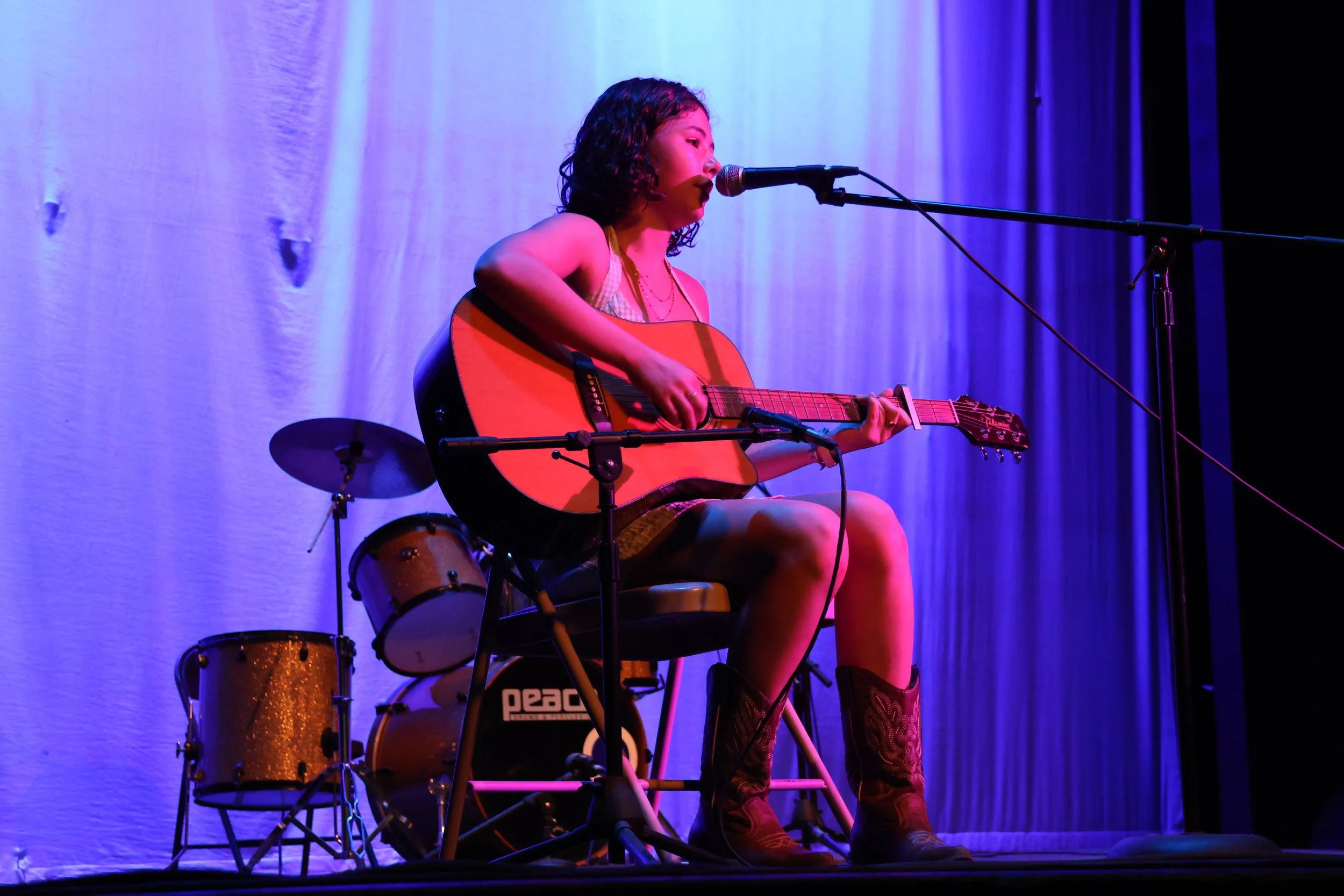 A woman with curly hair playing an acoustic guitar and singing into a microphone on a stage with purple and blue lighting. There are drums visible behind her.