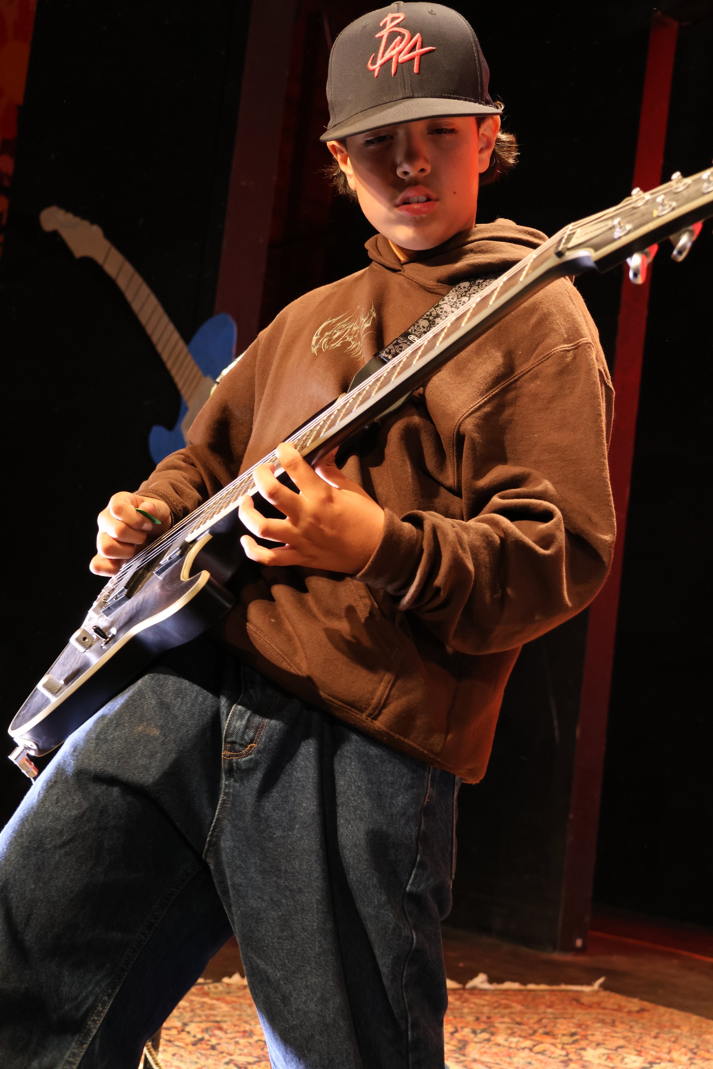 A teenage boy wearing a black baseball cap with red writing, a brown hoodie, and dark jeans, playing an electric guitar in a dimly lit room, possibly a stage or music studio.