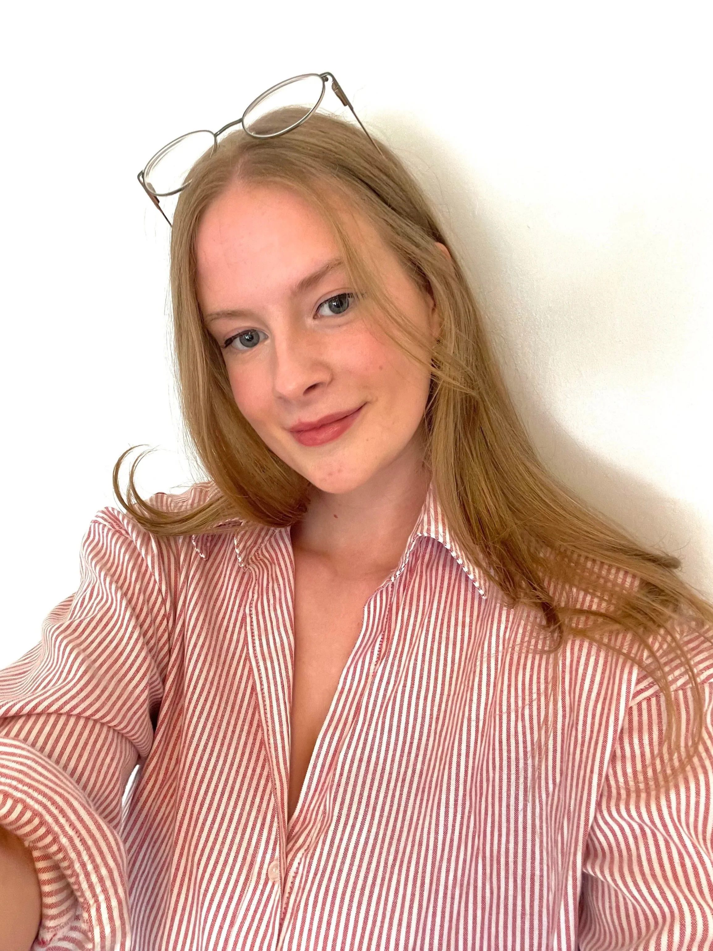 A woman with red hair and blue eyes wearing glasses on her head and a pink and white striped shirt, smiling softly against a plain white background.