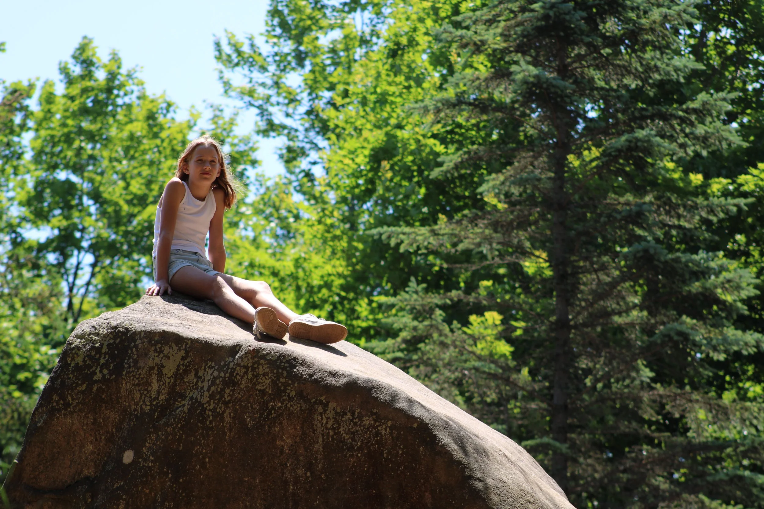 A young girl with light brown hair sitting on a large rock outdoors in a wooded area with green trees and blue sky in the background.