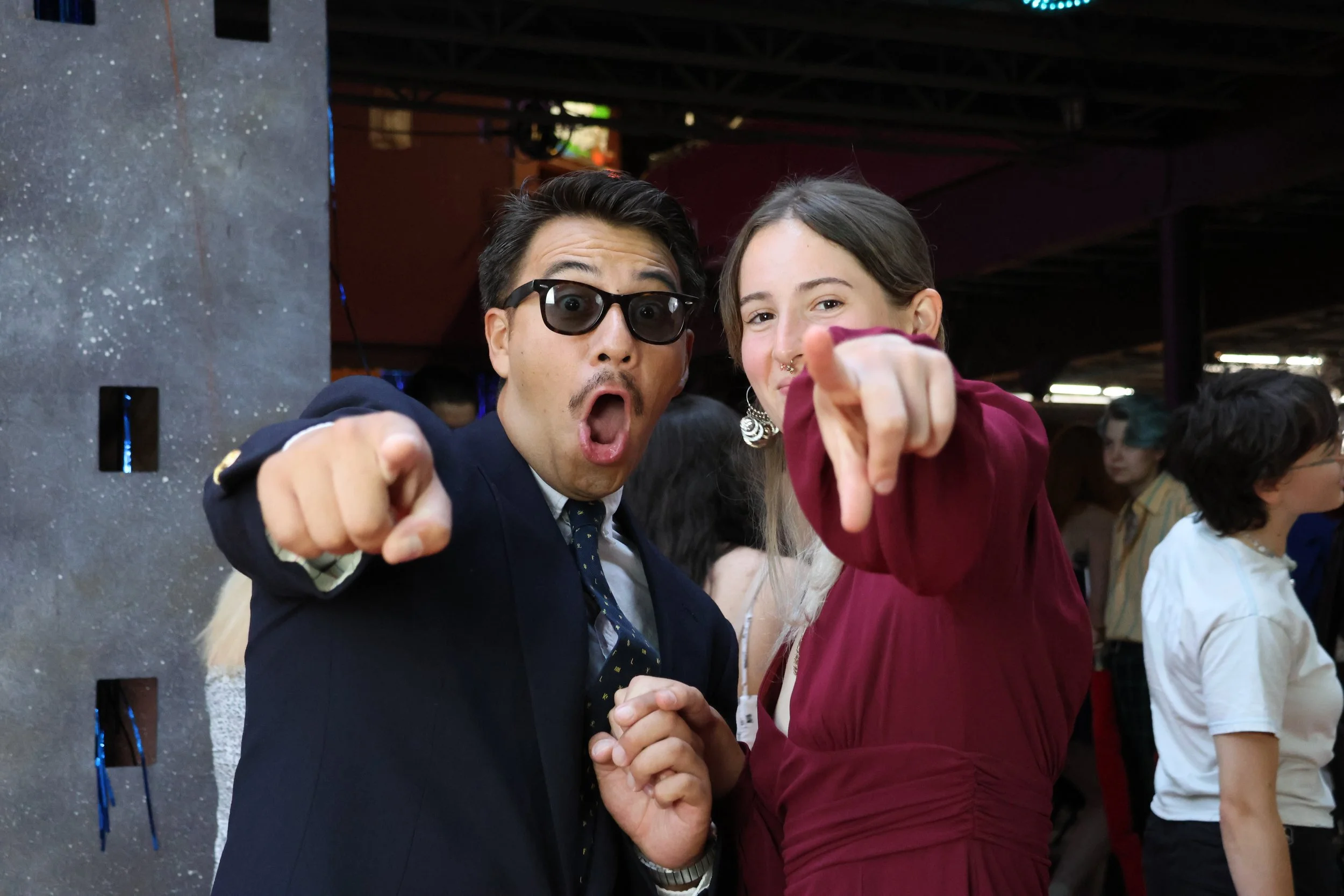 Two people, a man wearing sunglasses and a woman with a septum piercing, are engaged in a handshake with a playful, surprised expression. The woman points directly at the camera. They are indoors at an event with other people in the background.