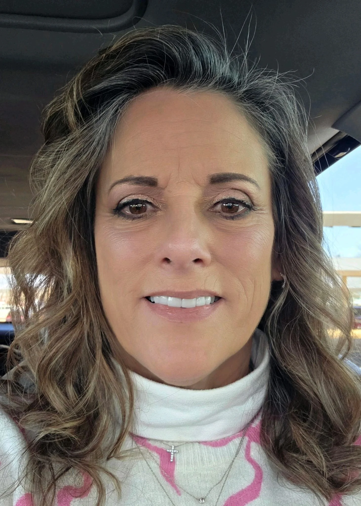 A close-up photo of a woman with wavy light brown hair with streaks of gray, wearing a white turtleneck with pink accents, smiling slightly, inside a car with a blue sky and parts of the outdoor surroundings visible through the windows.