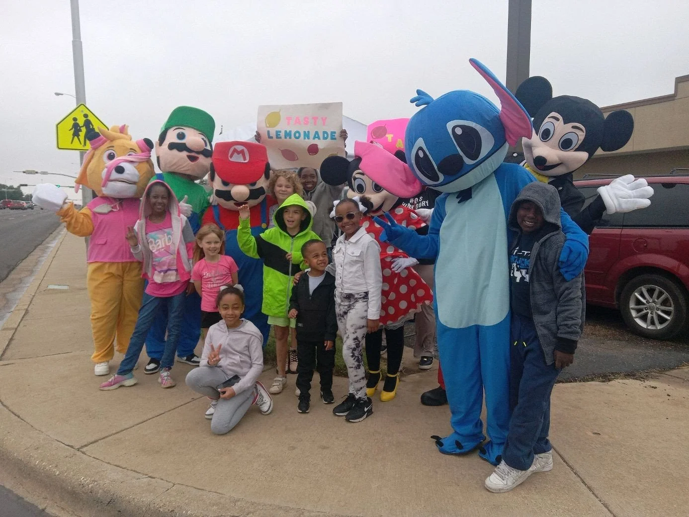Children posing with people in colorful costumes of cartoon characters including Mickey Mouse, Minnie Mouse, Stitch, and others during a parade or event.