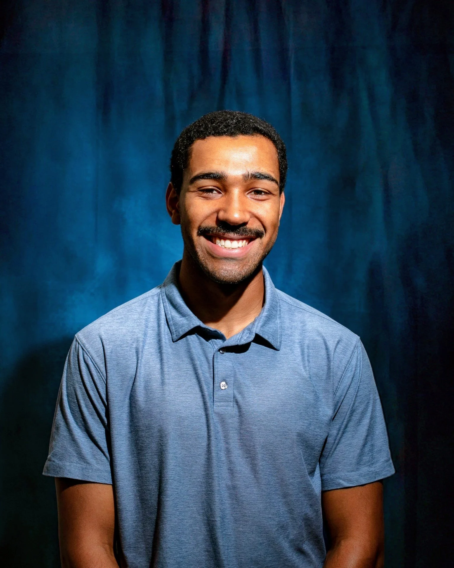 A smiling young man with dark curly hair and a mustache, wearing a light blue polo shirt, against a blue curtain background.