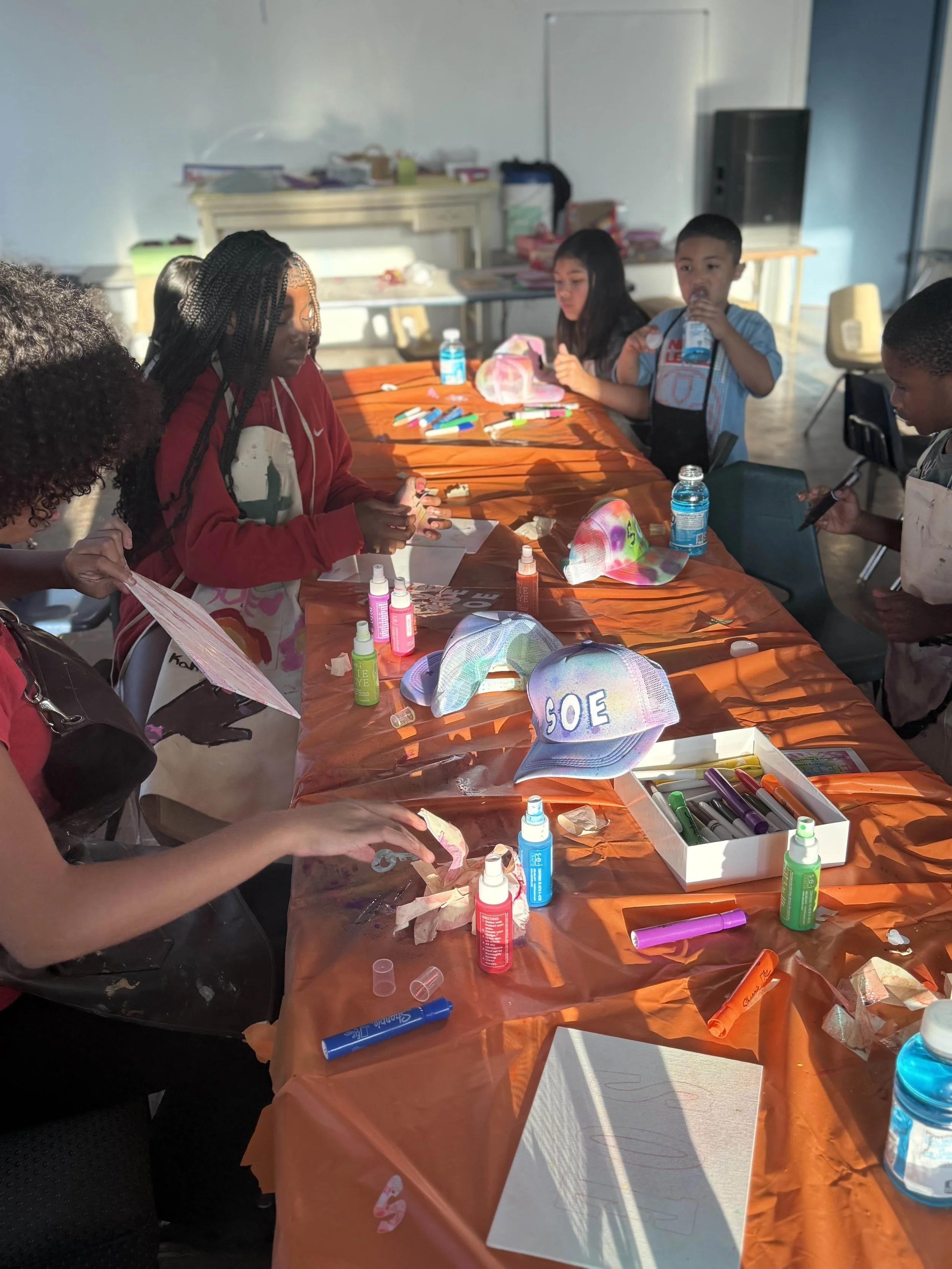 Children engaged in arts and crafts activity around a table covered with orange tablecloth, various craft supplies, and caps, with sunlight streaming into the room.