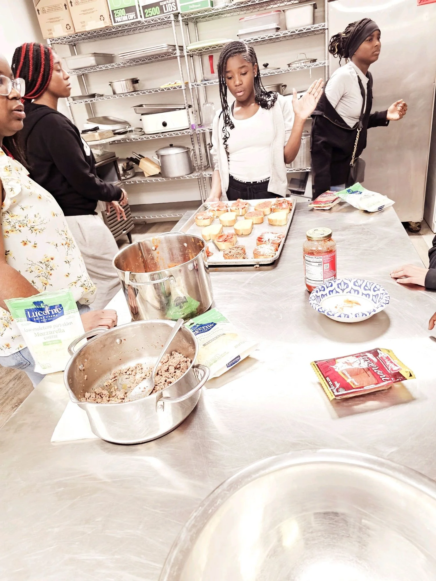 A group of children and adults in a kitchen or food preparation area, standing around a stainless steel table with food items, containers, and packages, some of which include cheese and ground meat. One girl is gesturing with her hand toward a tray of mini sandwiches or buns topped with various ingredients.