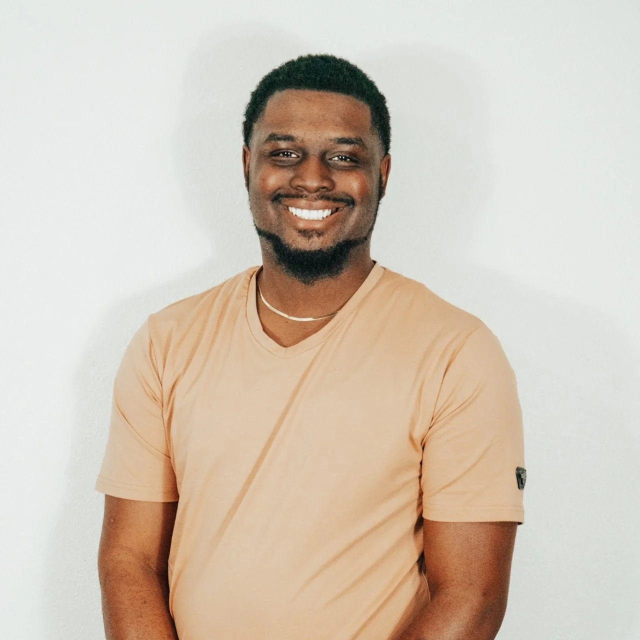 Smiling young man with dark hair, beard, and mustache wearing a beige t-shirt and a necklace, standing against a white wall.