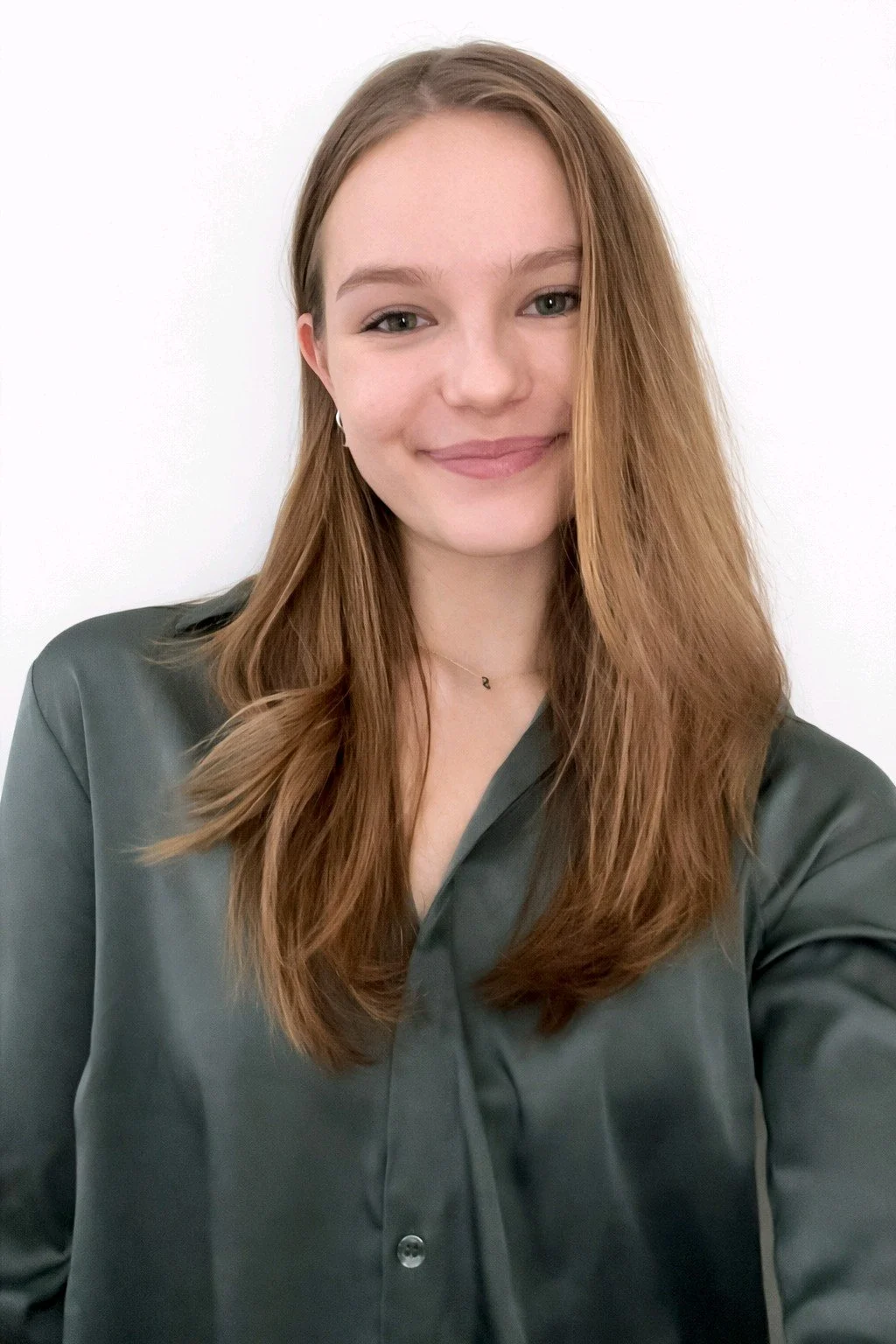 A young woman with long, light brown hair, smiling, wearing a dark gray button-up shirt and small earrings, against a plain white background.