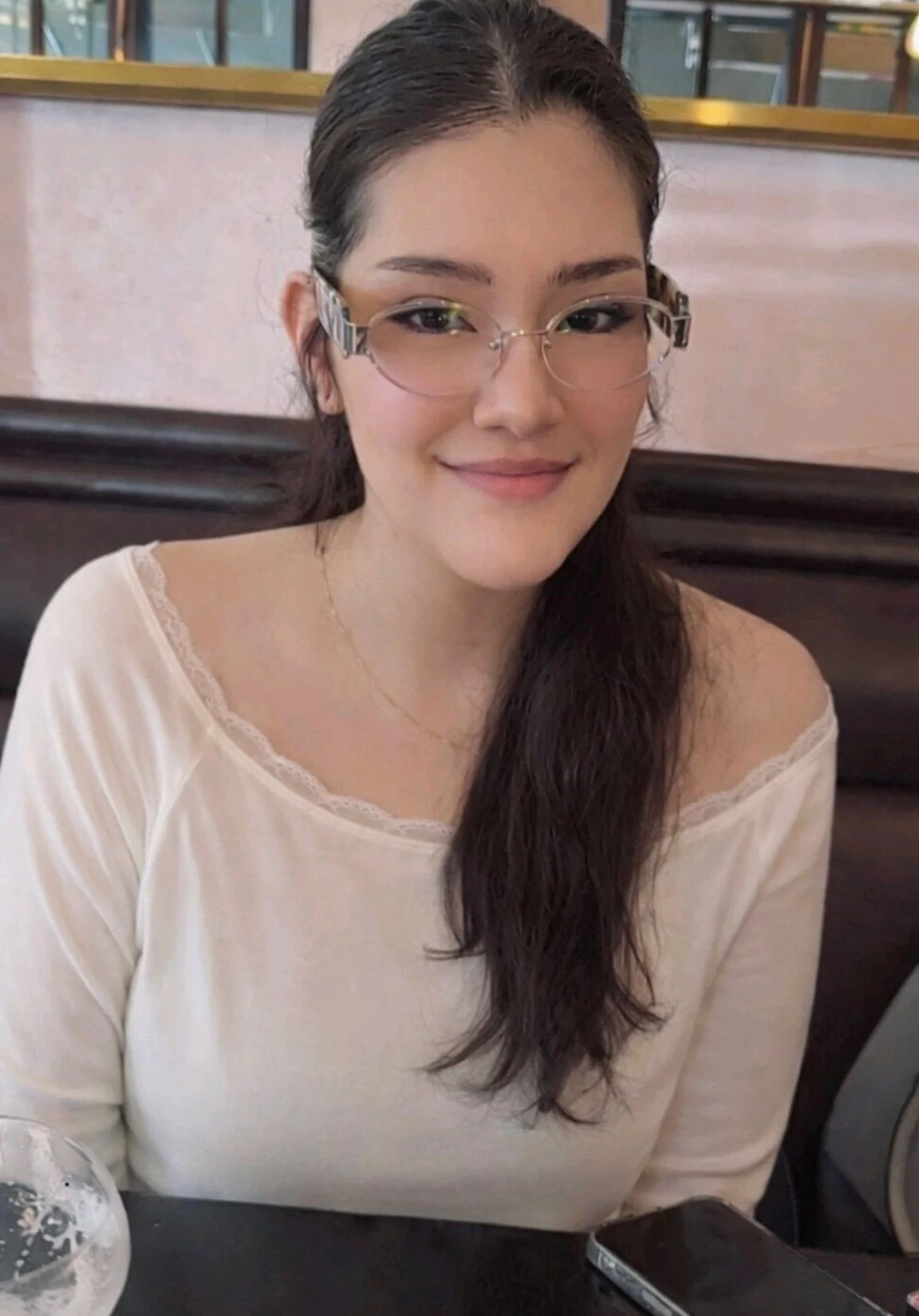 A young woman with long, dark hair, wearing glasses and a cream-colored top, sitting at a table in a cafe or restaurant.