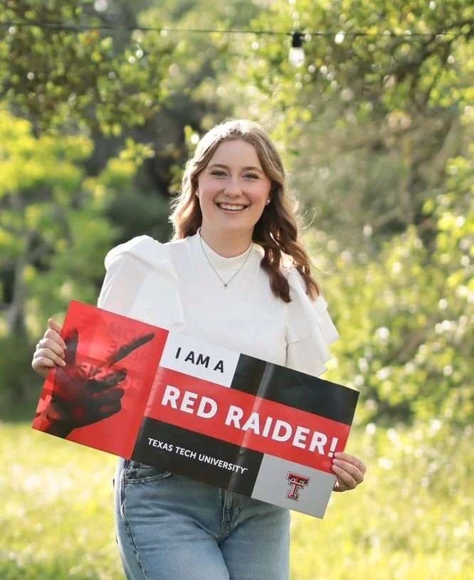 A young woman smiling outdoors holding a Texas Tech University sign that says 'I am a Red Raider!' with green trees and sunlight in the background.