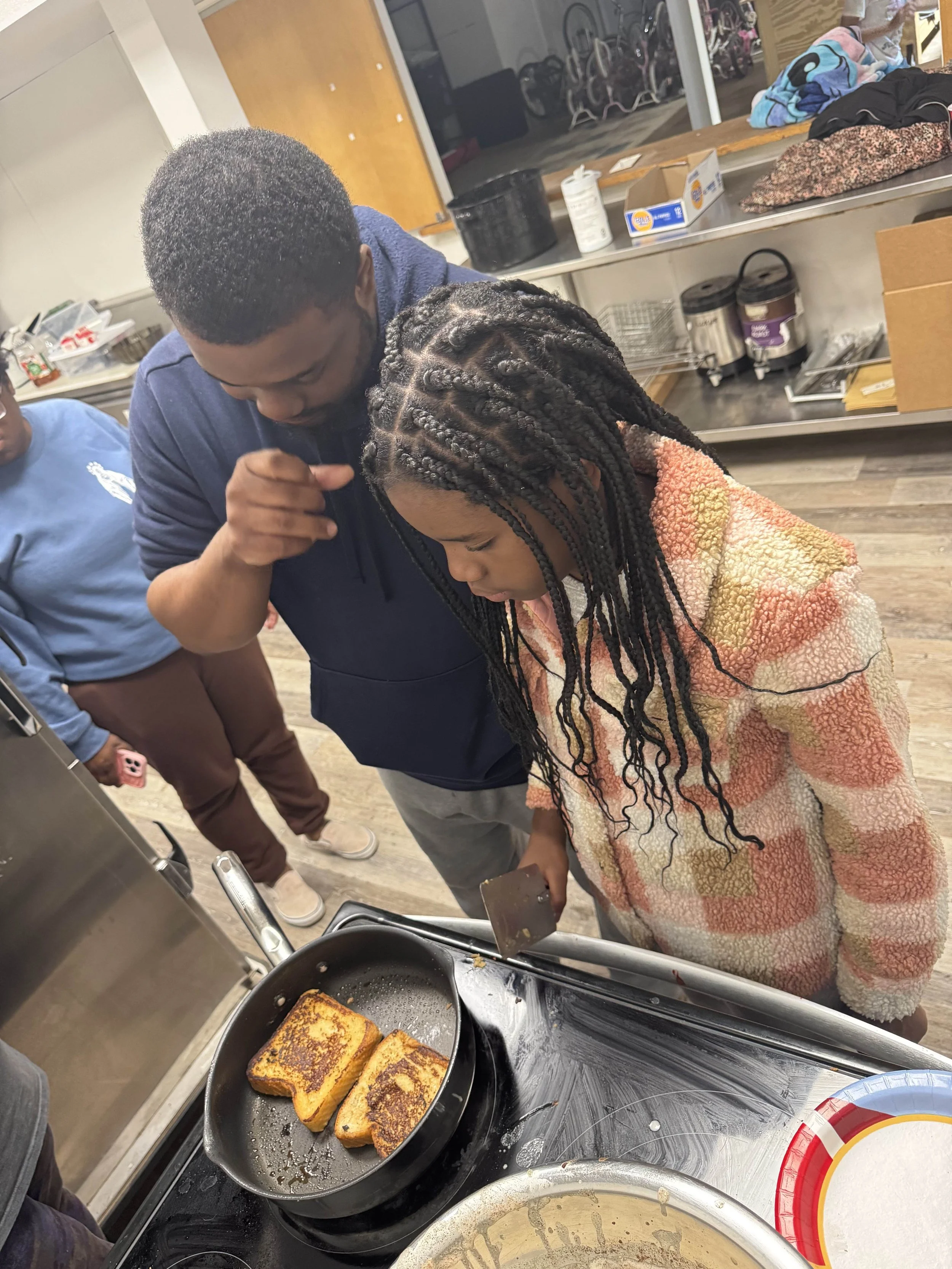 A man with short hair and a young girl with braids are cooking toast in a kitchen. The man is assisting the girl as she holds a spatula and looks at the toast. There is a frying pan with toasted bread, a stove, and various kitchen items in the background.