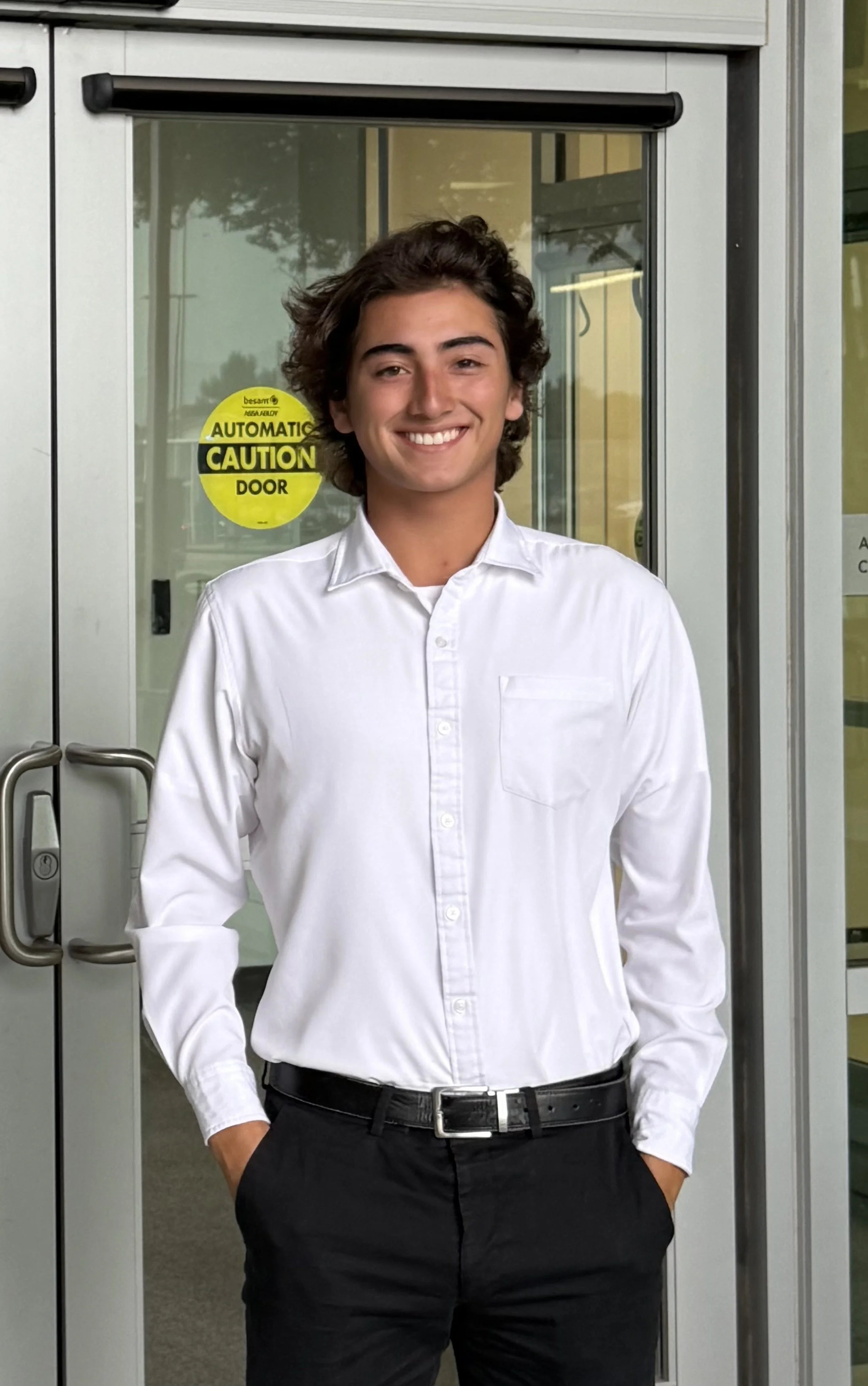 Young man standing in front of glass door, smiling, wearing a white dress shirt and black pants.