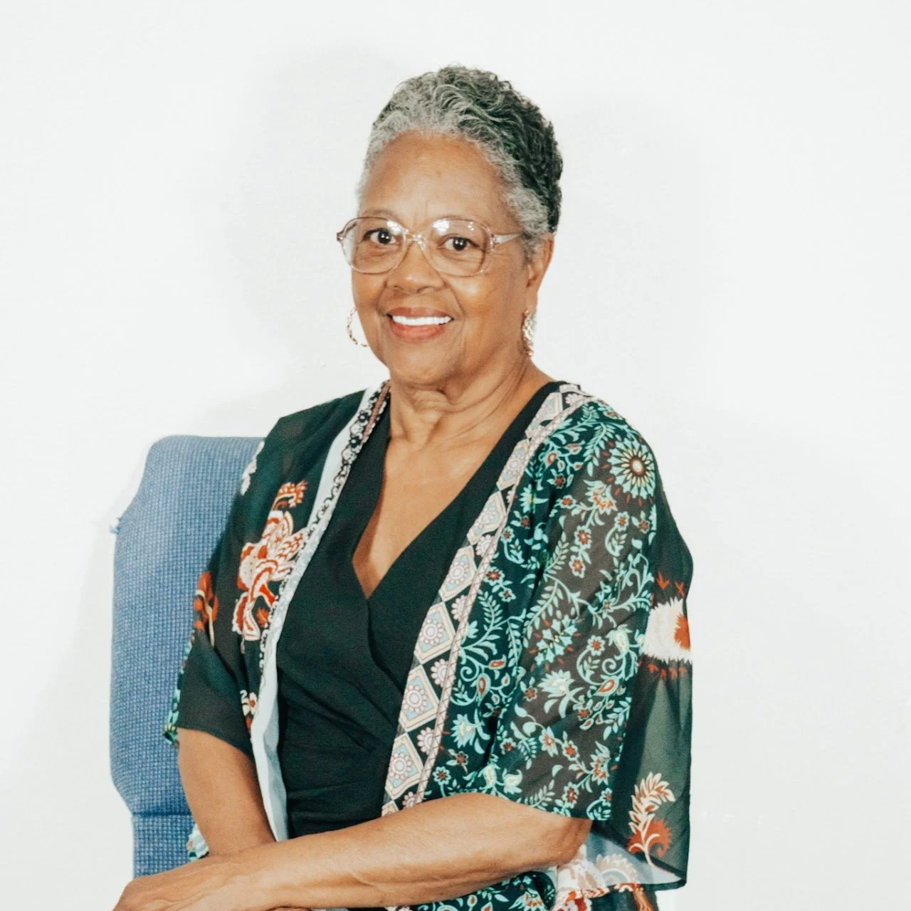 An elderly African American woman with short gray hair, glasses, and earrings, smiling and sitting in a blue chair against a white background.