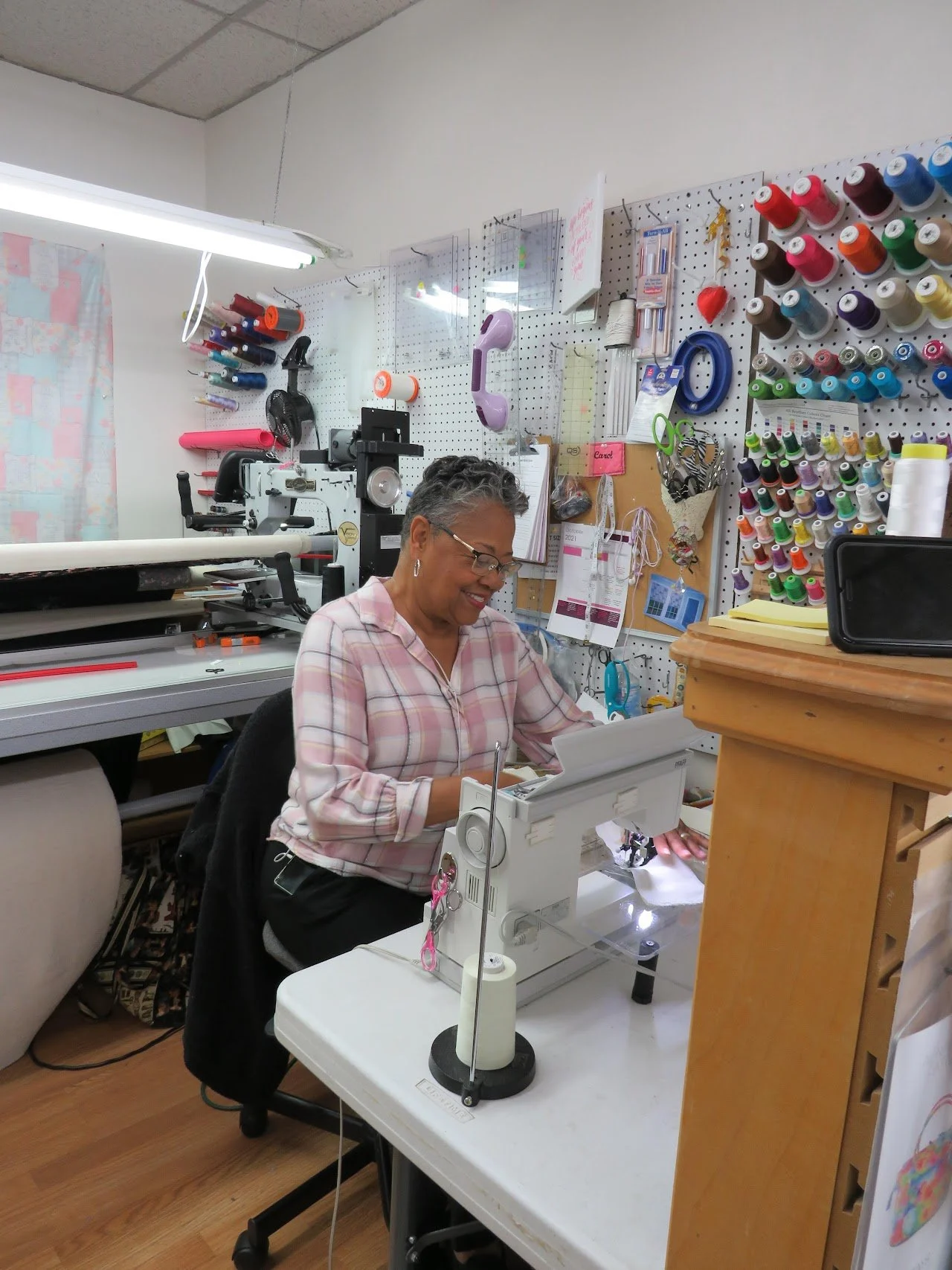 A woman sitting at a sewing machine in a sewing or craft room, surrounded by thread spools, fabric, and sewing tools.