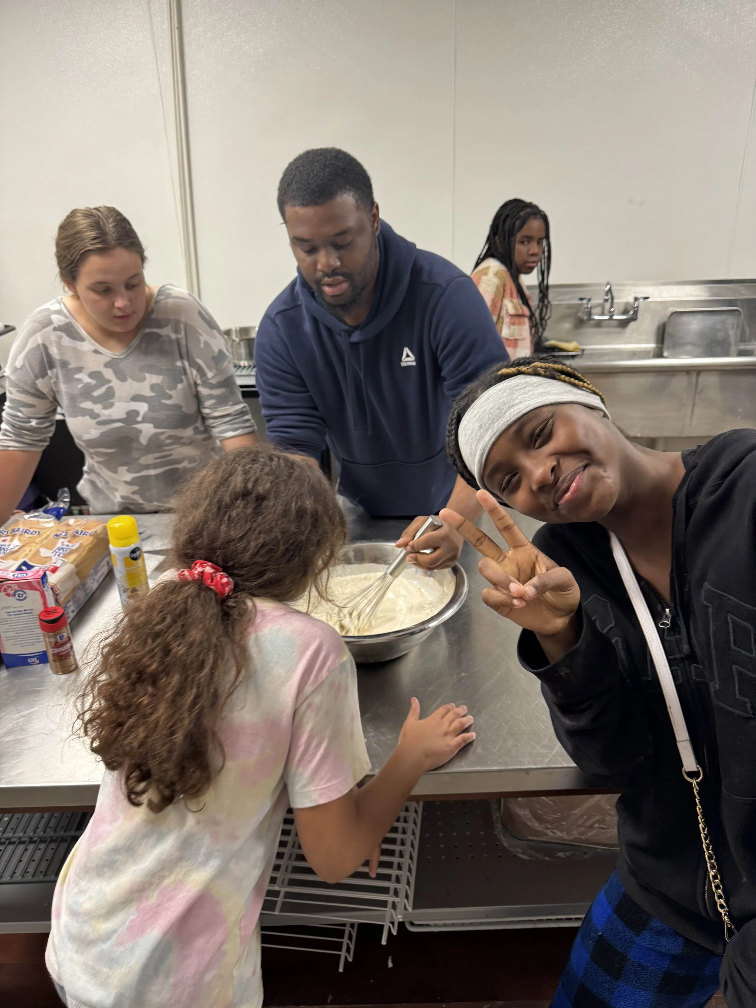 Group of people cooking together in a kitchen, one person making a peace sign and smiling.