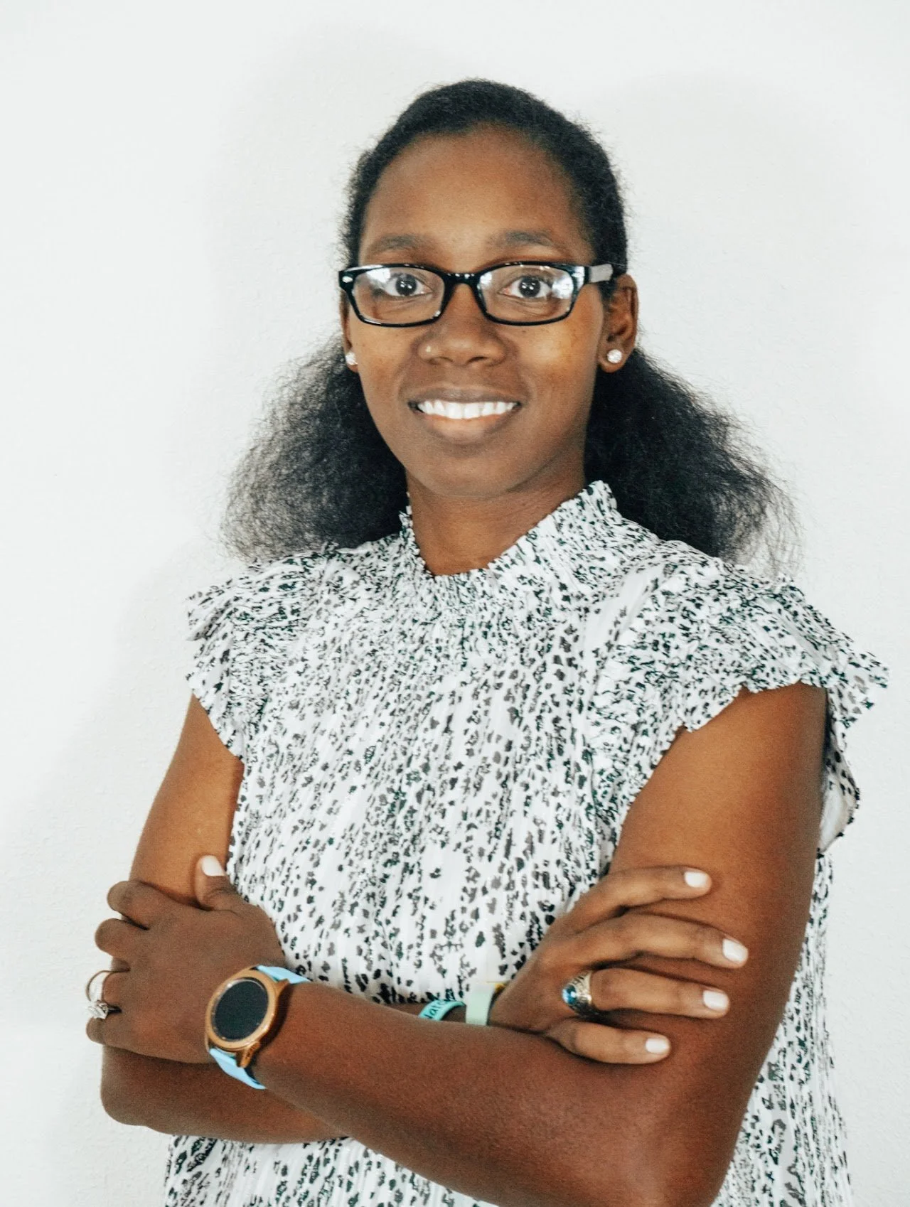 A woman with glasses and earrings, wearing a patterned sleeveless top, standing with arms crossed in front of a plain white wall.