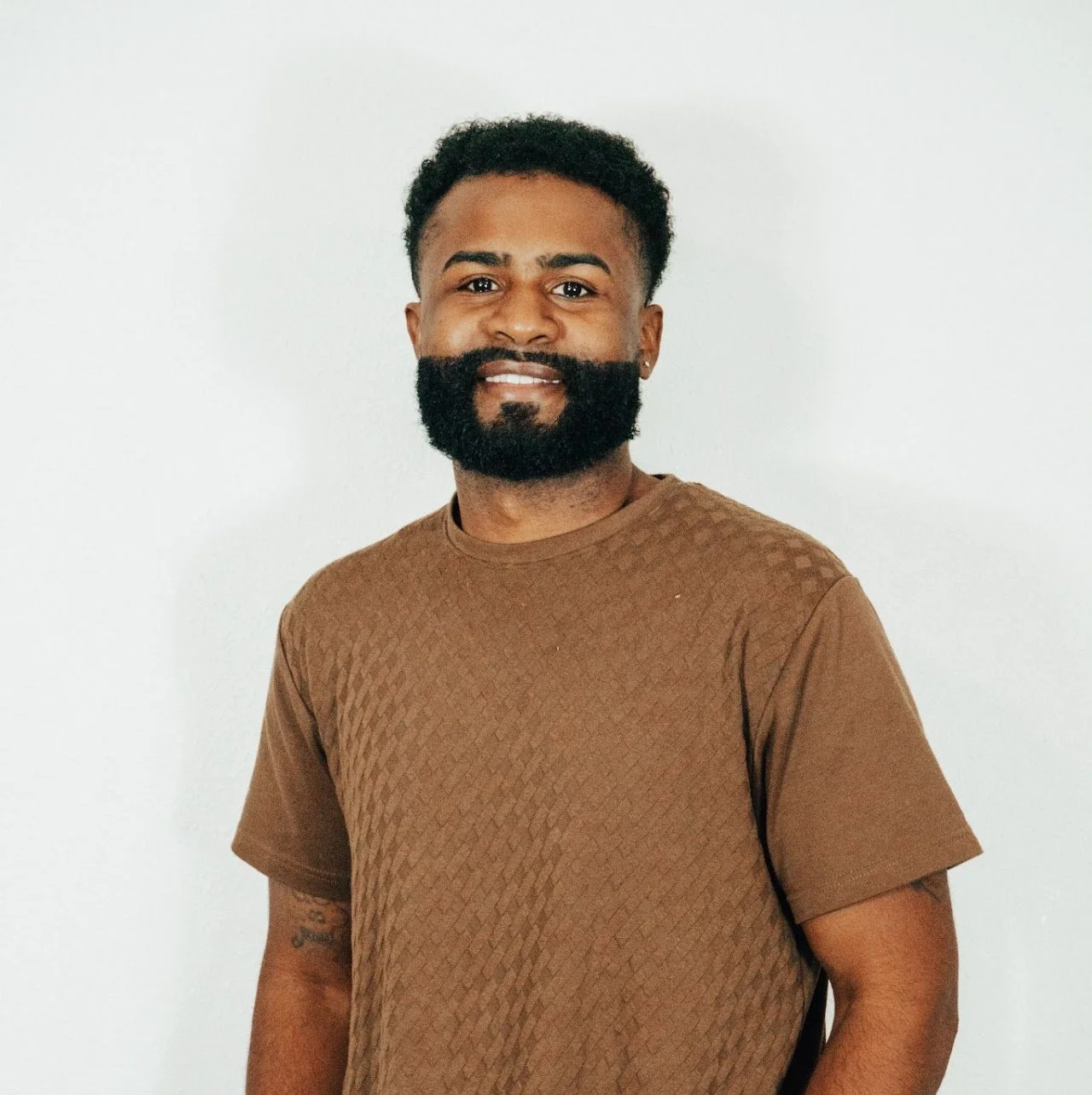 A man with a beard and short curly hair smiling, wearing a brown textured t-shirt, standing against a plain white background.