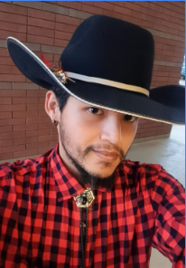 A person wearing a large black cowboy hat with a white band, a red and black checkered shirt, and a bolo tie, standing in front of a brick wall.