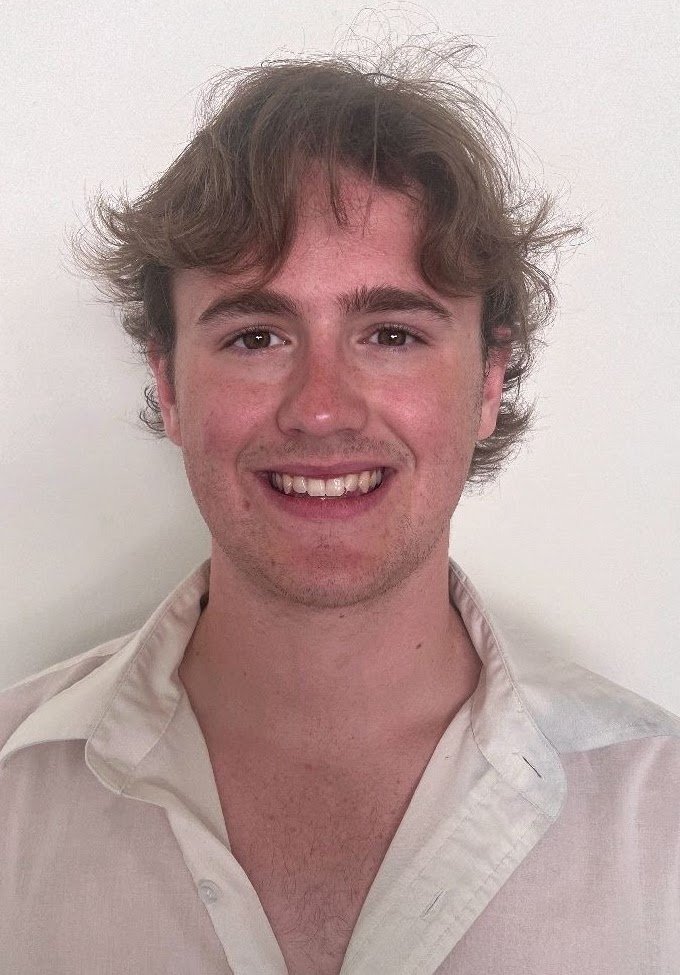 A young man with wavy brown hair and light skin, smiling, wearing a cream-colored shirt against a plain white background.
