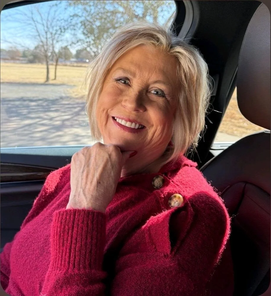 A smiling older woman with blond hair in a red sweater sitting in a car, looking out the window with a scenic outdoor view in the background.