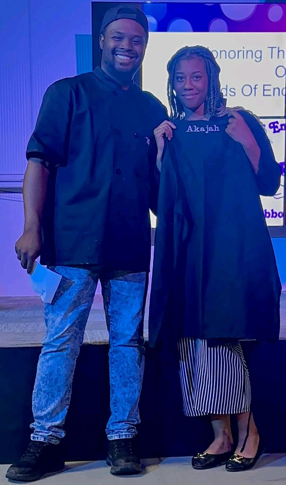 A young woman in a graduation gown and cap holding a diploma, standing next to a man in a black chef's coat, in front of a screen at an indoor event.